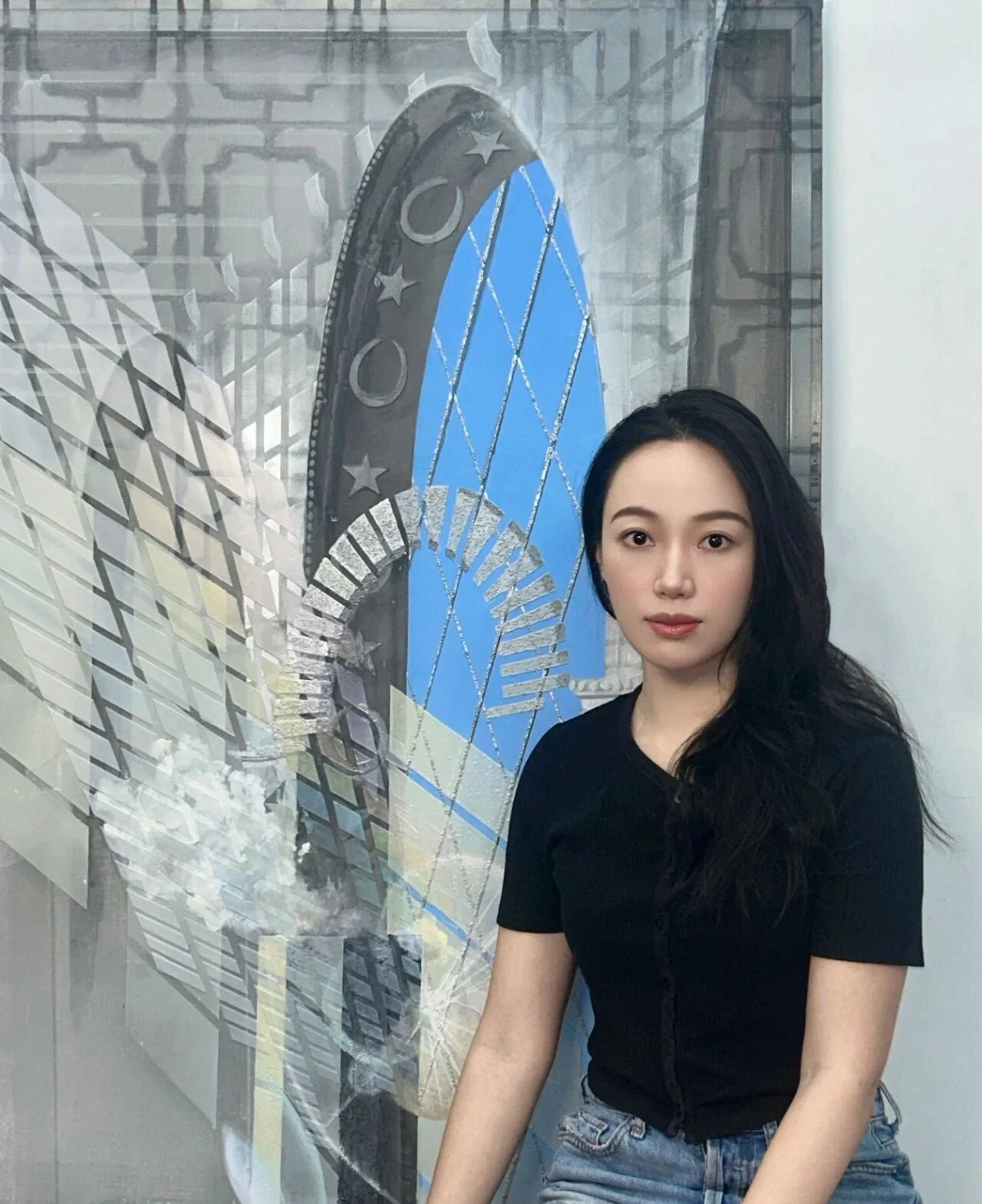 Young Asian woman with long black hair wearing a black shirt and jeans standing indoors in front of a wall art with a blue and black geometric design.