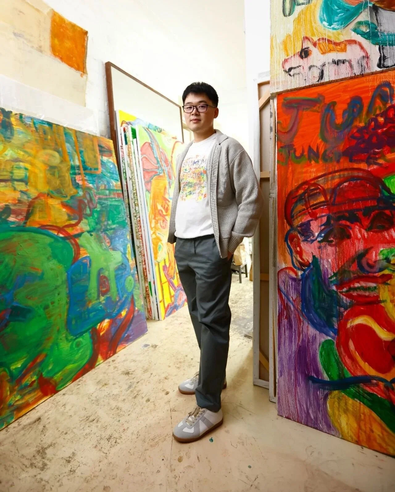 A young man stands with hands in pockets among colorful abstract paintings in an art studio.