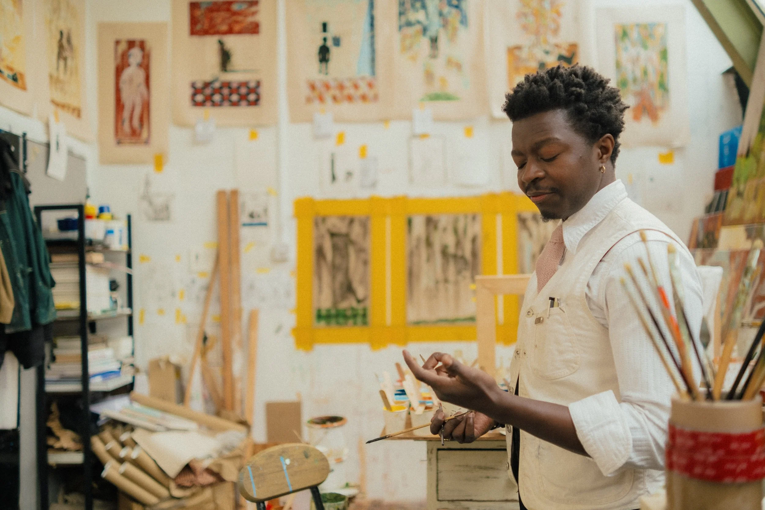 A man with curly hair and earrings in an art studio, holding a paintbrush, standing near an easel with artwork. The background features walls decorated with colorful paintings and art supplies on shelves.