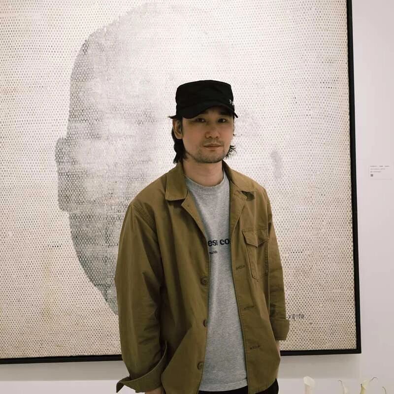 A young man with dark hair and facial hair, wearing a black cap, a gray t-shirt, and a brown jacket, standing in front of a pixelated portrait artwork in an art gallery.
