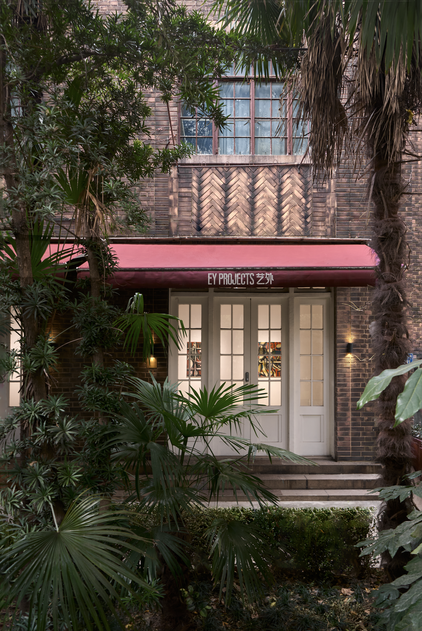 A brick building with a door and a window above it, partially obscured by lush green plants and trees.