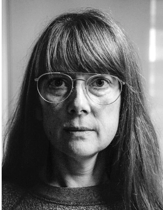 A black and white photo of a woman with long hair, wearing round glasses, looking directly at the camera.