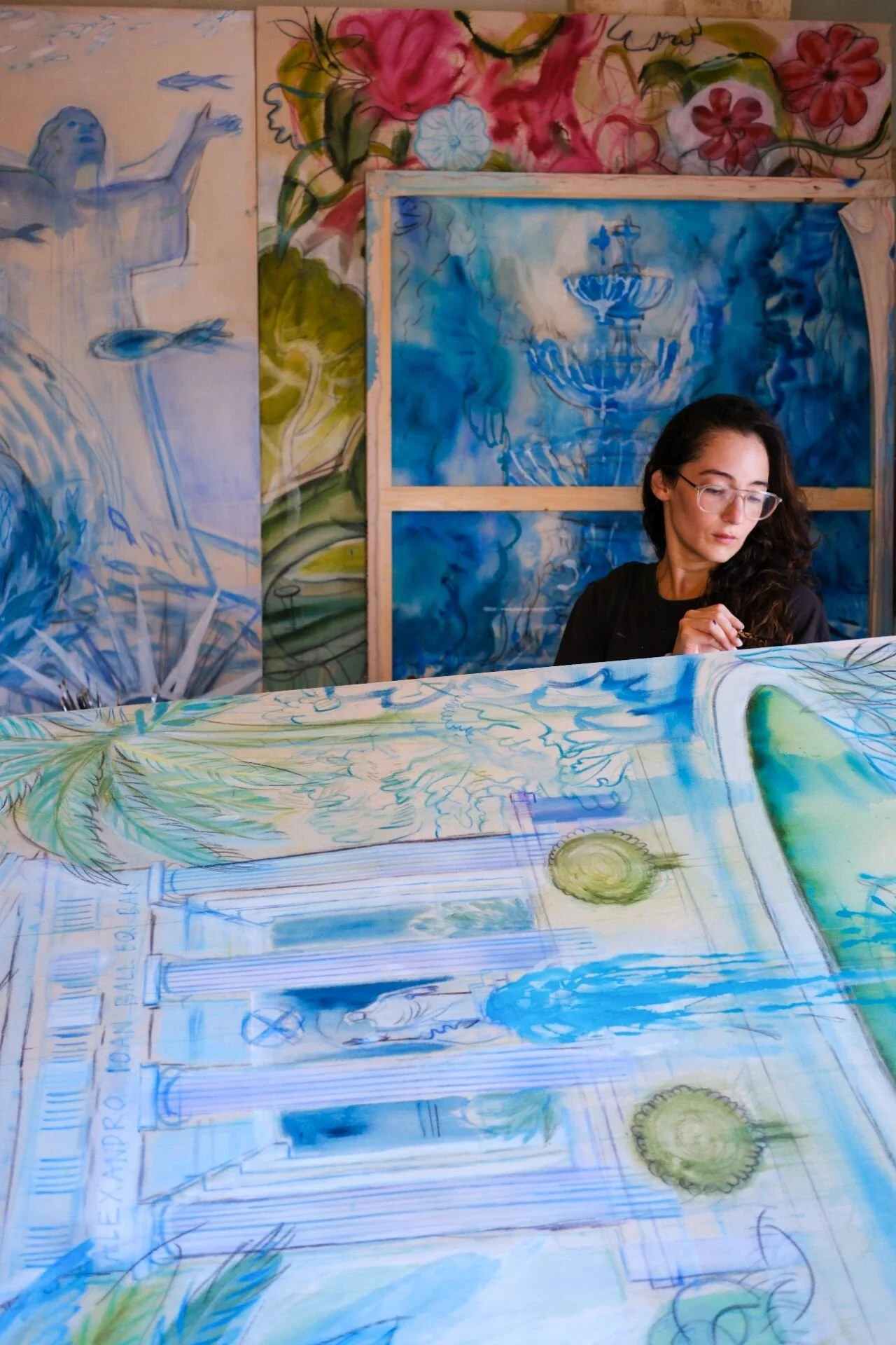 An artist sitting in front of a large tabletop covered with sketches and watercolor paintings of a building and landscape scenes. Behind her, there are large painted panels of water fountains, flowers, and greenery.