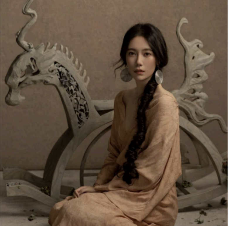Young woman with long braided hair and large earrings sitting in front of an ornate, carved wooden deer sculpture.