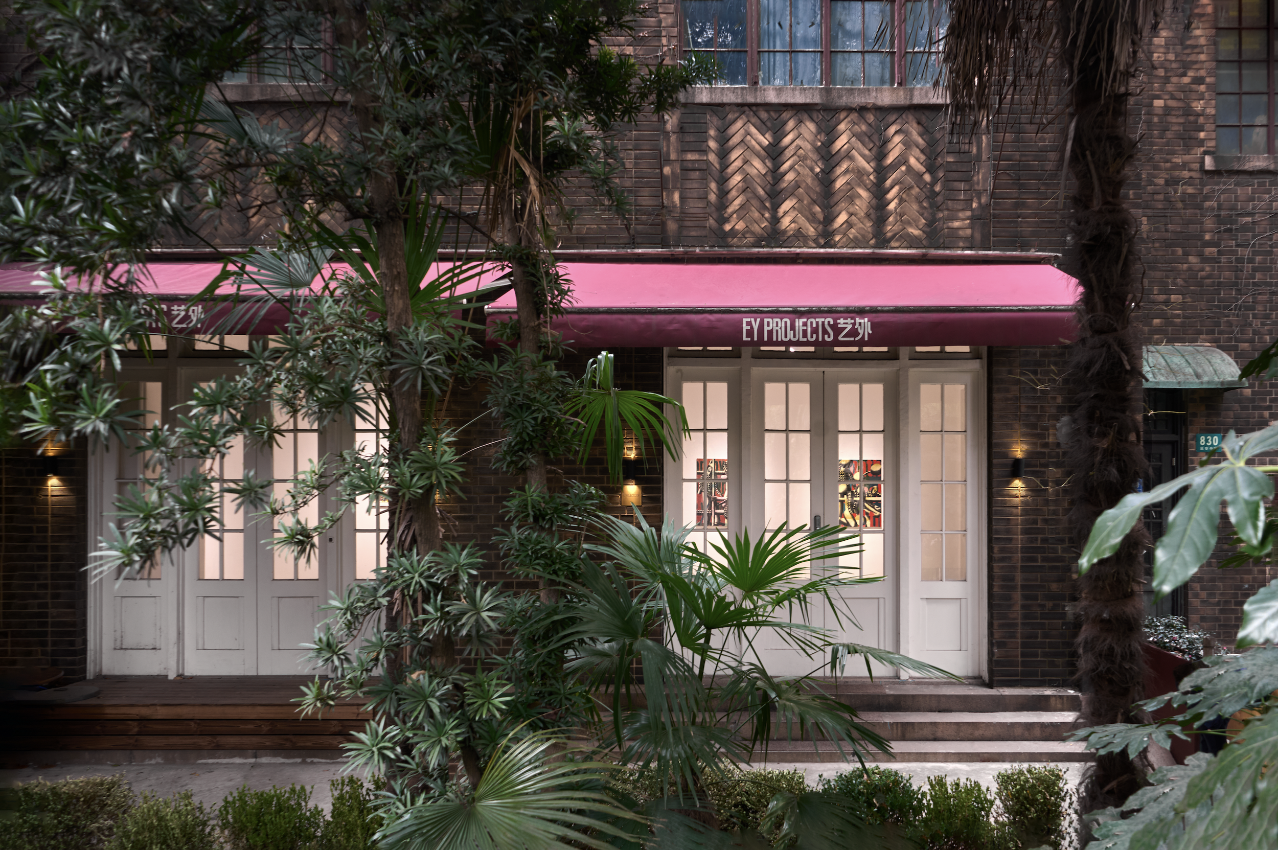 The image shows the exterior of a building with a red awning that has the text "EY PROJECTS" on it. The building walls are made of dark bricks, and there are multiple windows on the upper floor. The scene is partially obscured by green plants and trees in front of the building.
