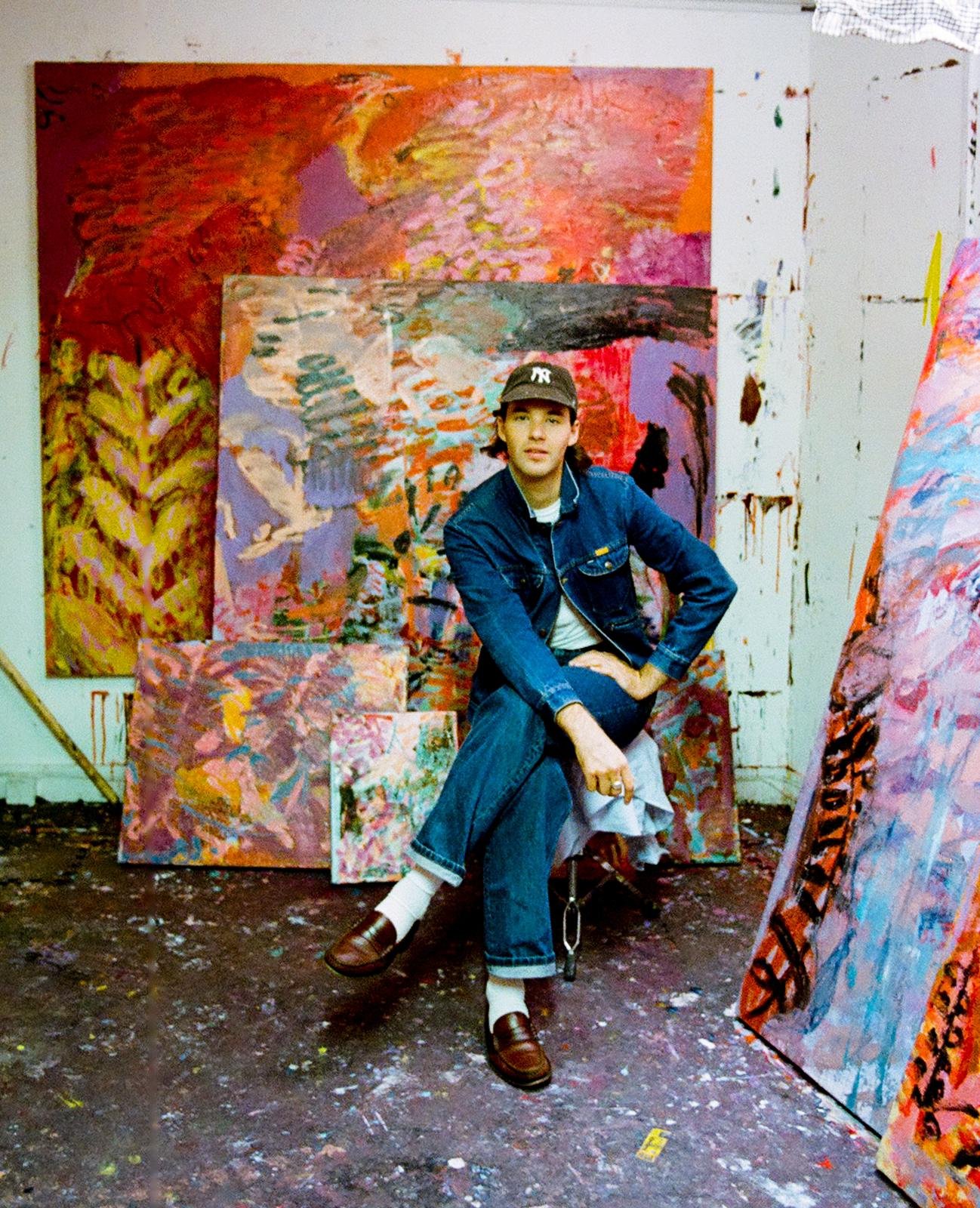 A man in a denim jacket and dark jeans with rolled-up cuffs, sitting in an art studio surrounded by colorful abstract paintings.