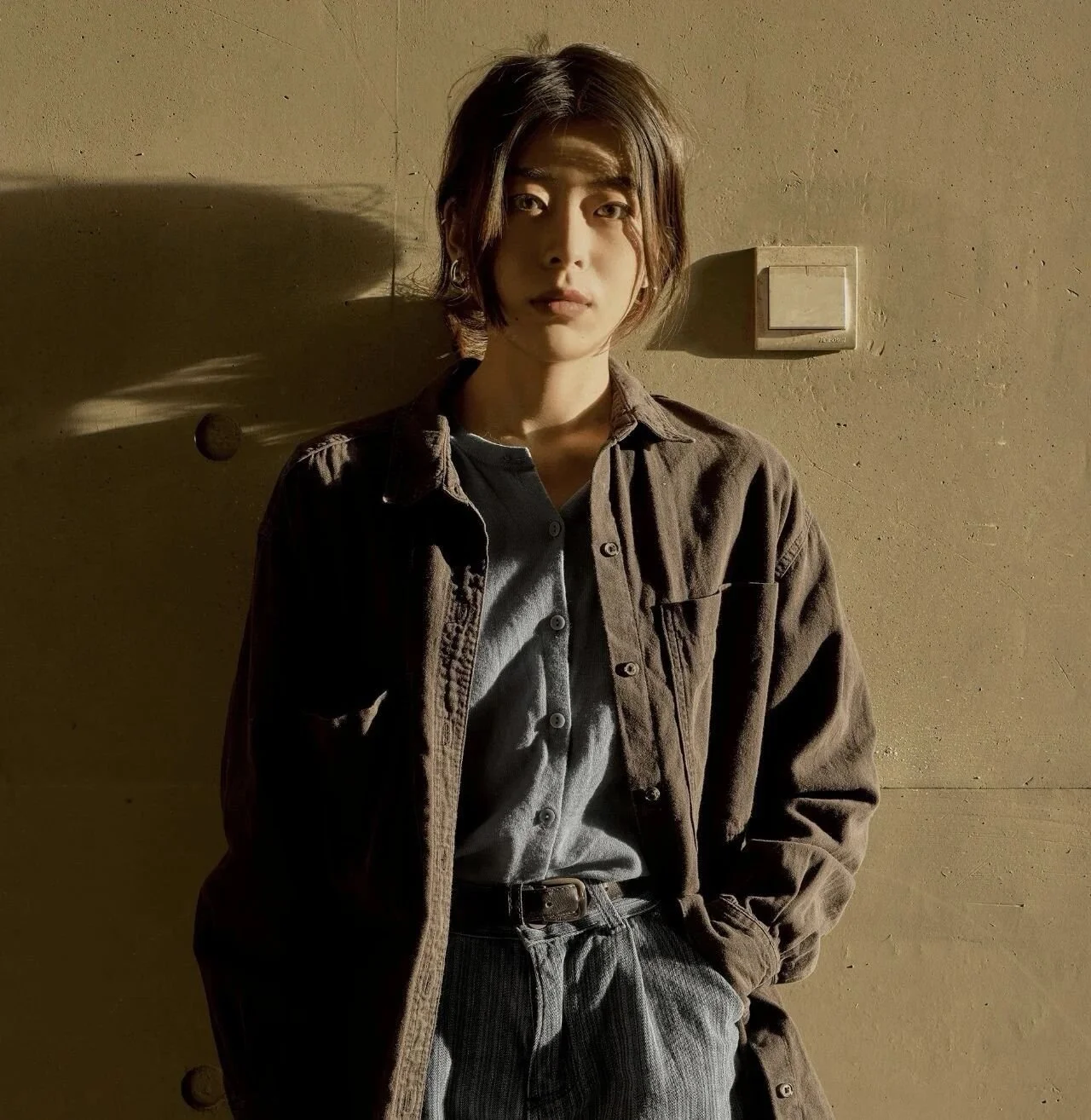 A young woman with short, dark hair standing against a beige wall, with sunlight casting shadows on her face and clothing.