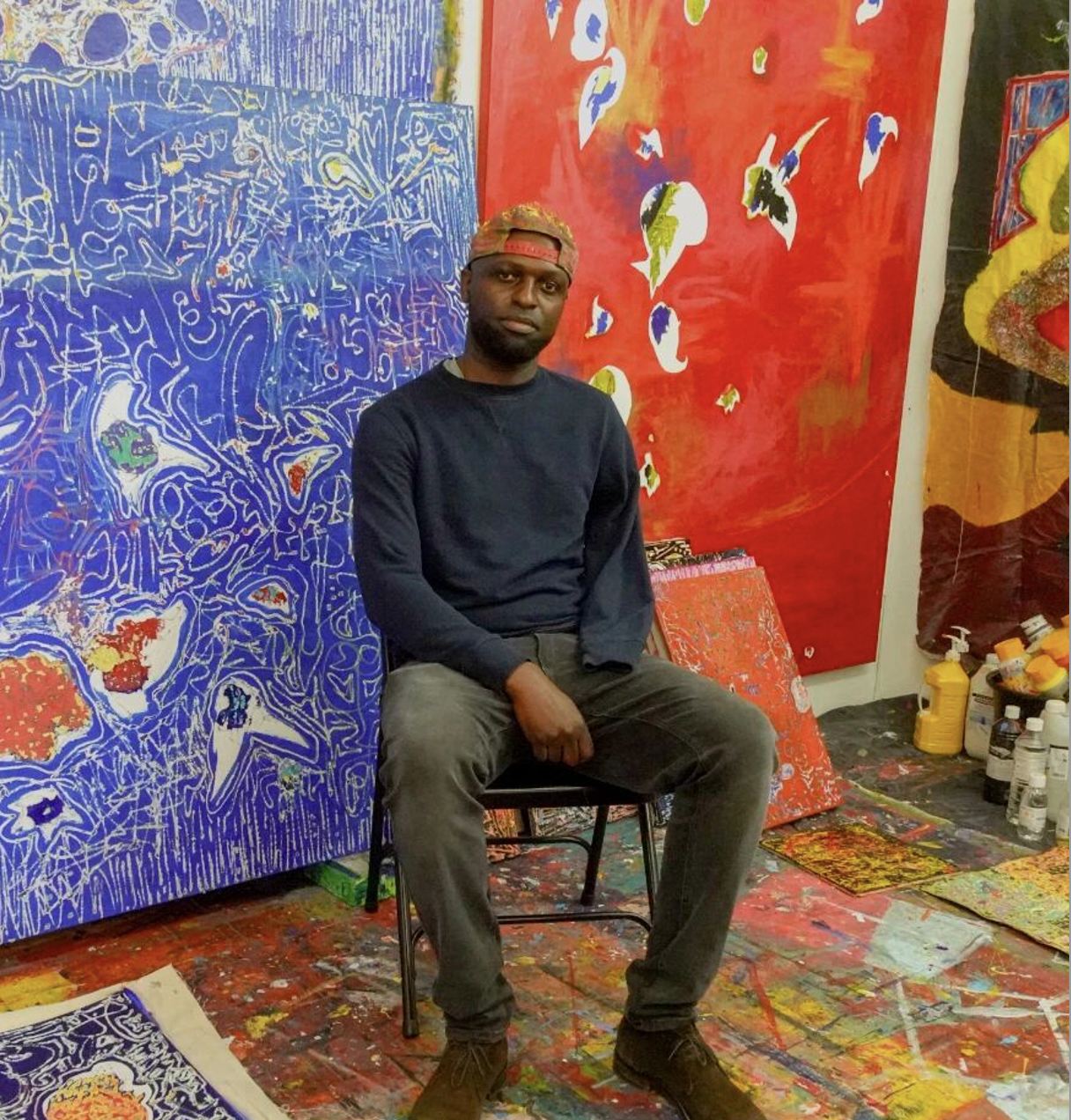 A man sitting on a black chair in an art studio surrounded by colorful abstract paintings and art supplies.