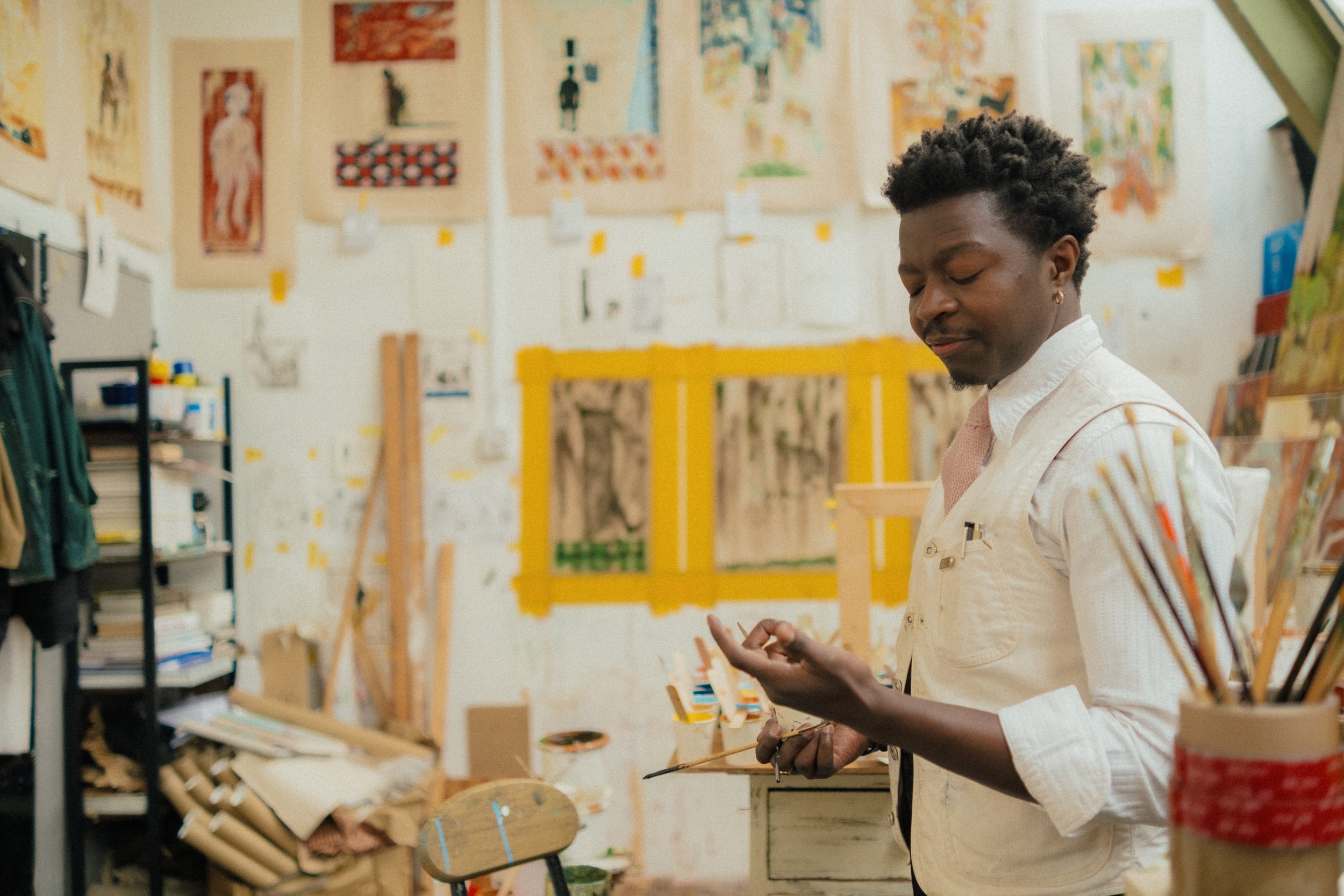 Person with short curly hair wearing a beige shirt, standing in front of an art display in an art studio or gallery.