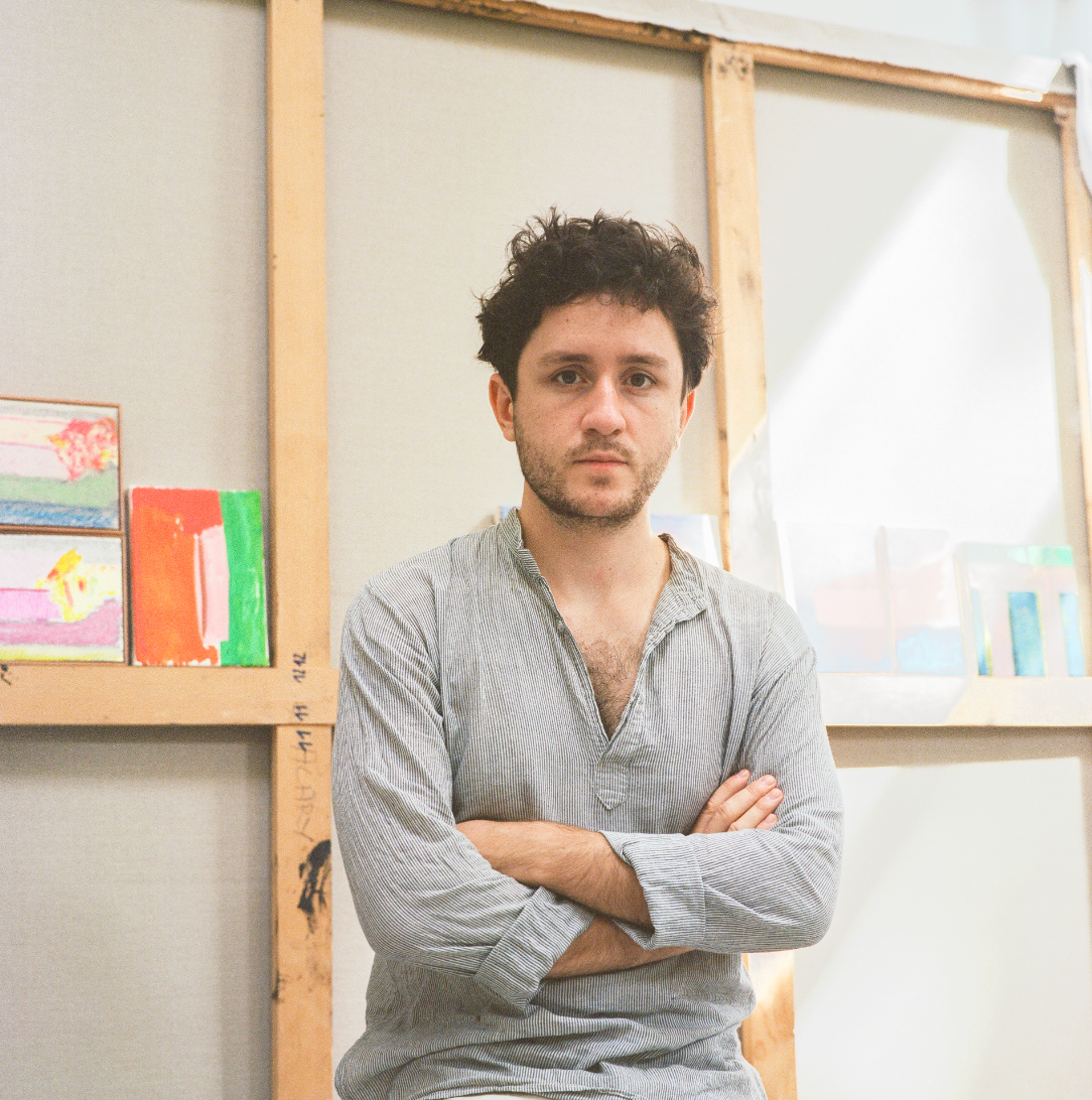 A young man with curly dark hair and a beard, wearing a gray striped button-up shirt with rolled-up sleeves, standing with arms crossed in an art studio with unfinished wooden frames and colorful artworks in the background.