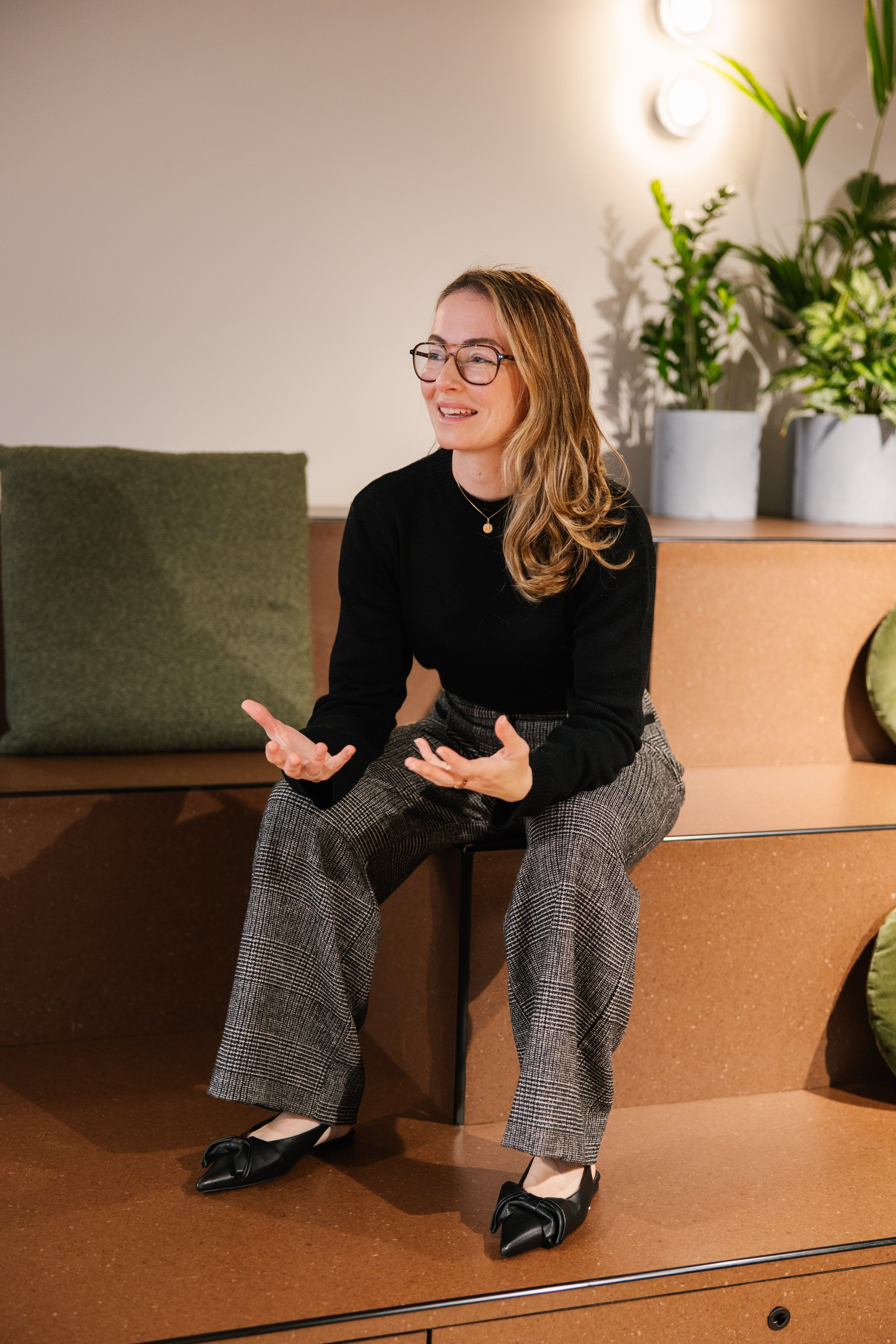 A woman with glasses and long wavy hair, wearing a black sweater and checkered pants, sitting on a chair and talking, with a plant and light fixtures in the background.