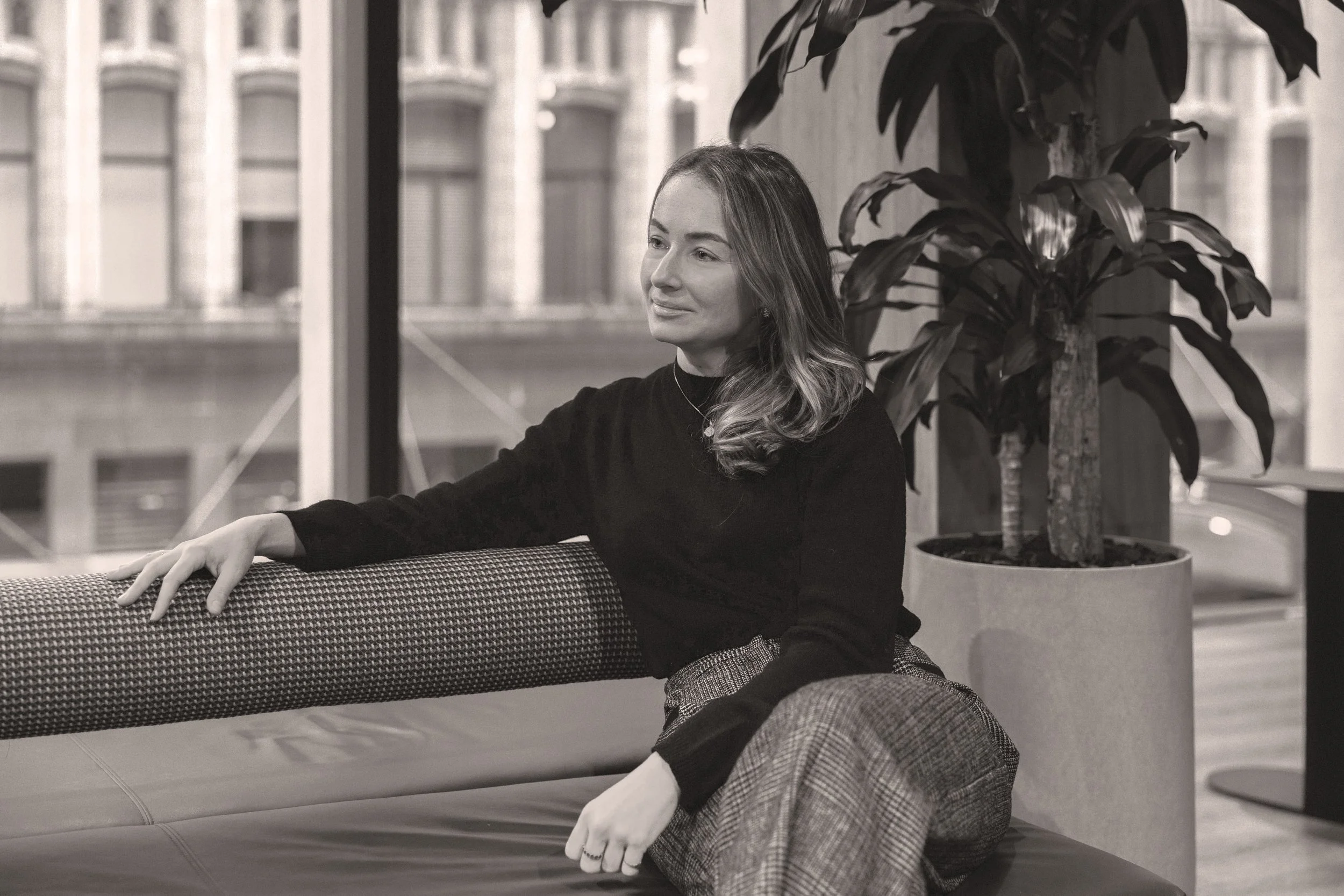 A woman with wavy hair sitting on a couch, smiling, with a large potted plant behind her, indoors with a cityscape visible through the window.