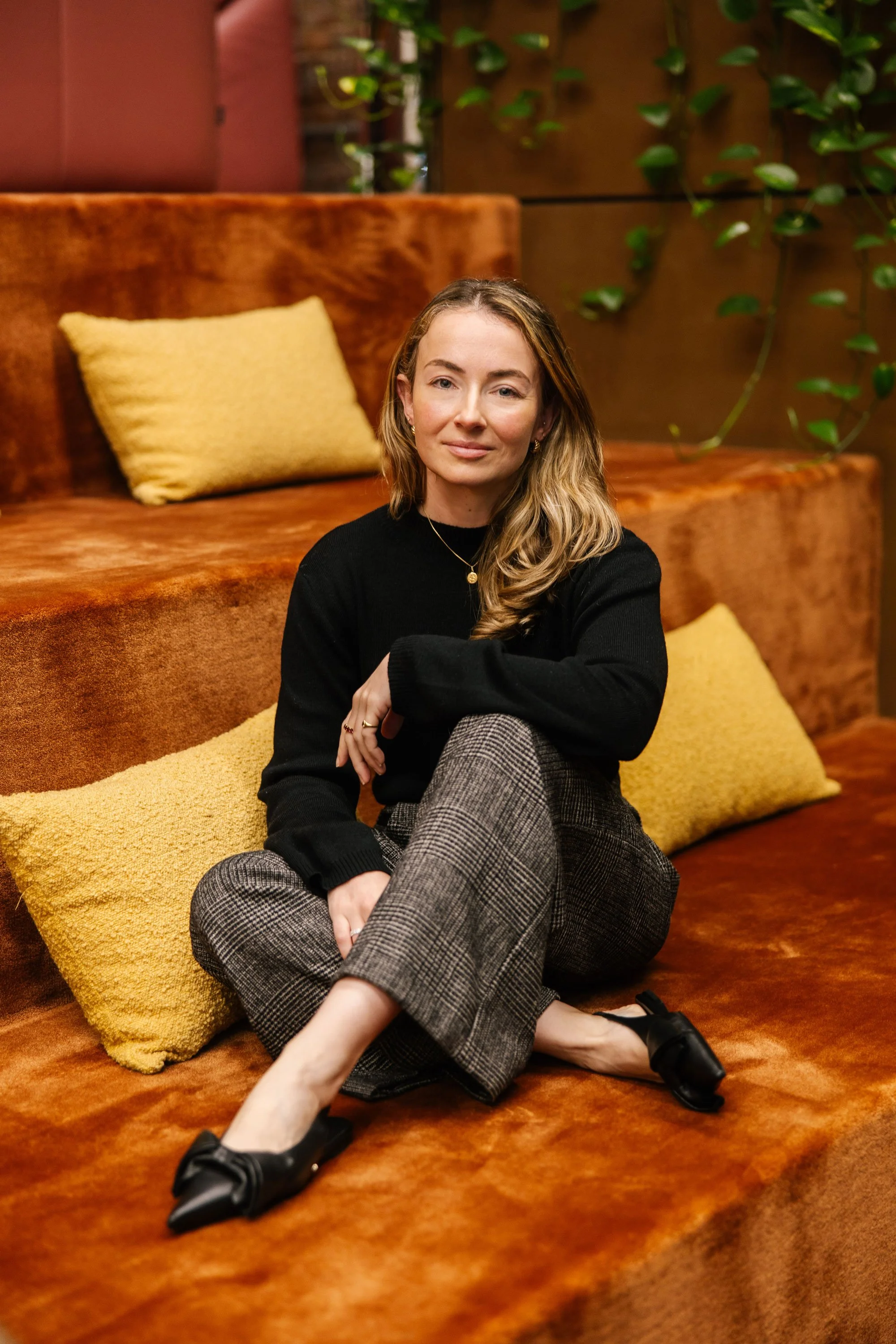 A woman sitting on a burnt orange sofa with a yellow pillow behind her. She is wearing a black sweater and checkered pants, with one leg crossed over the other, and black shoes. The background features a brown wall with green vines.