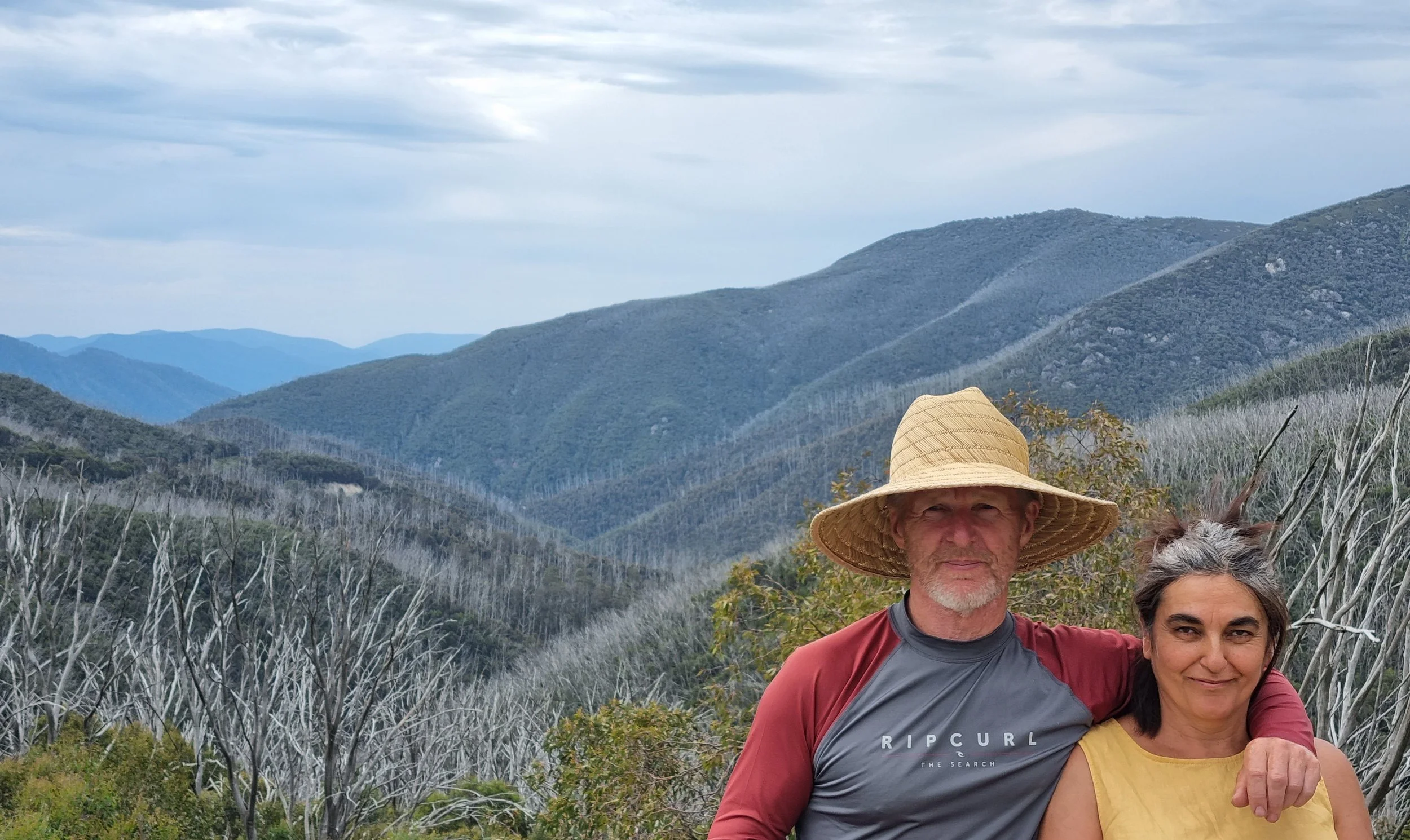 A man and woman standing outdoors in a mountainous area with cloudy skies, some dry vegetation, and a hat worn by the man.