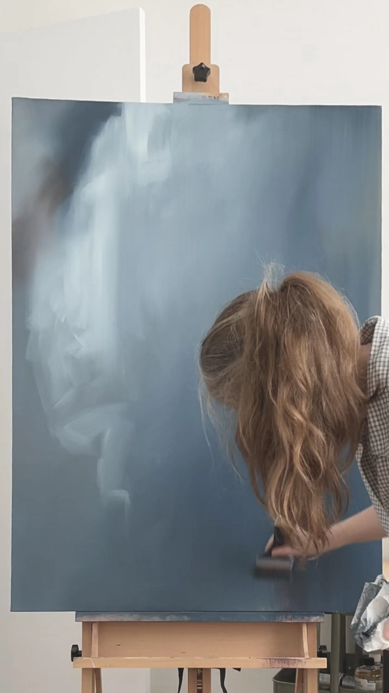 A woman with long red hair is painting on a blue canvas, focusing on creating a gradient effect. The person is leaning over, side view, wearing a checkered shirt.