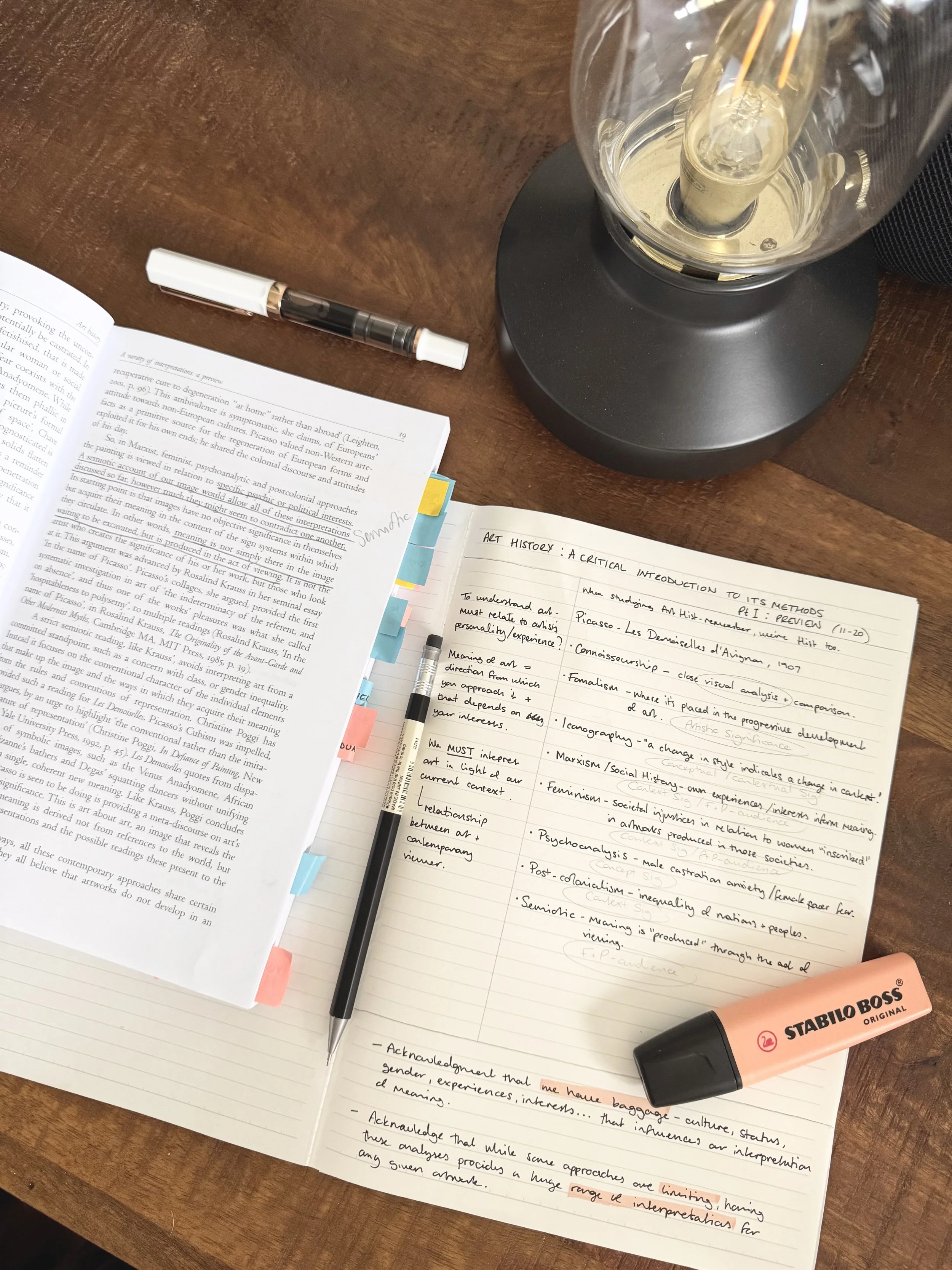 A wooden desktop with open notebooks, colorful sticky tabs, a pen, a pencil, a peach highlighter, and a black table lamp. The notebooks contain handwritten notes and printed text.