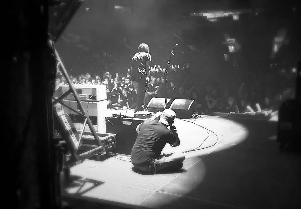 Black and white photograph of a performer on stage playing an electric guitar, with a seated photographer capturing the moment, audience in the background