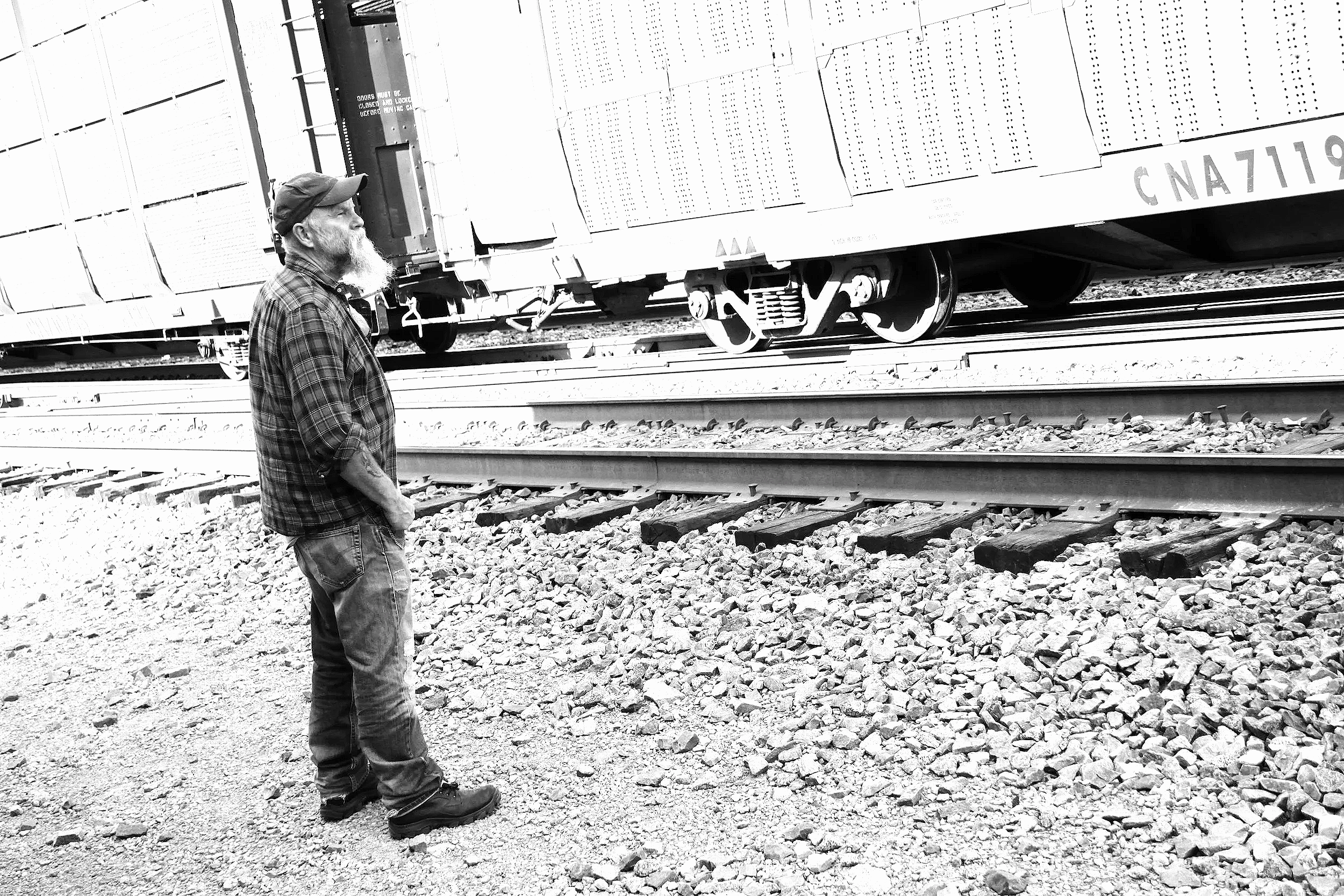 Seasick Steve wearing a plaid shirt, jeans, and a cap, standing near a freight train car on railway tracks in Nashville, Tennessee.