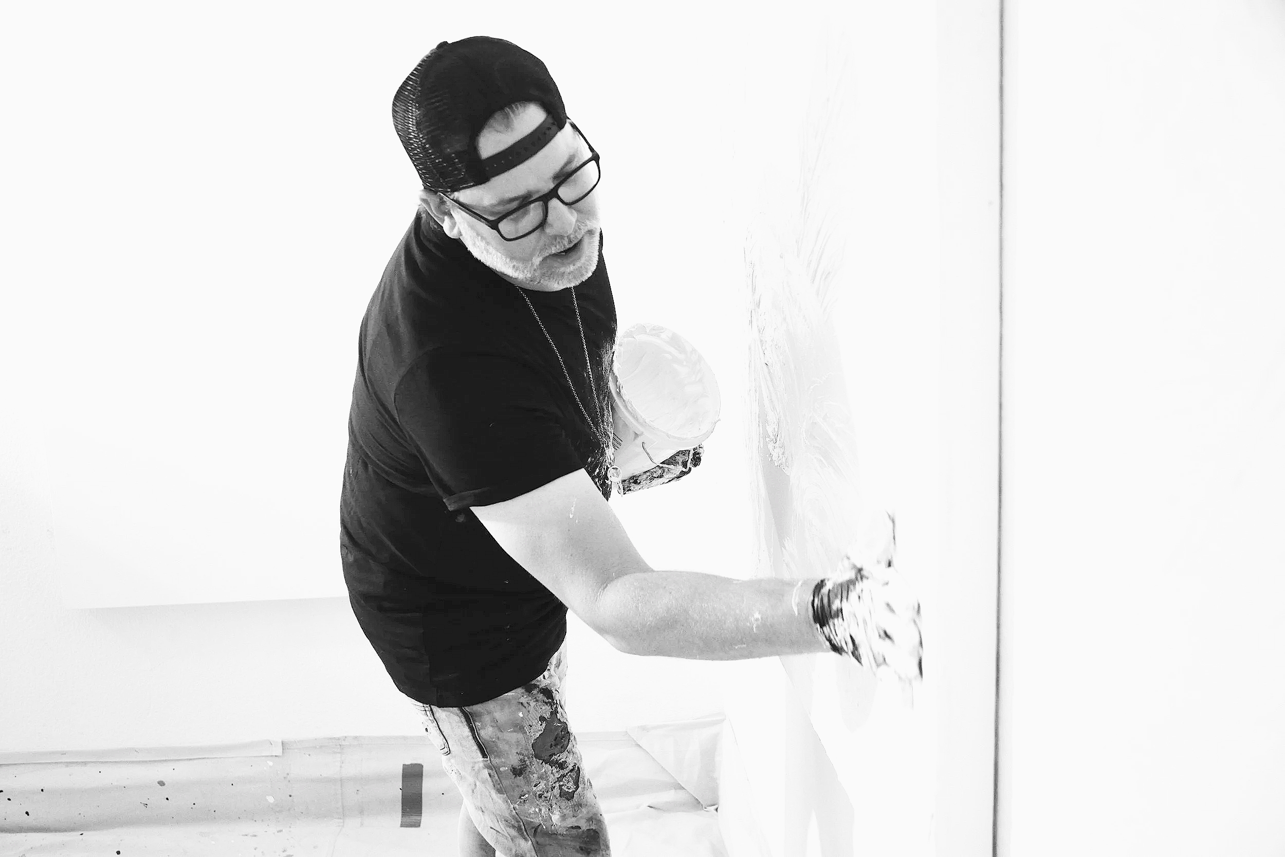 A black-and-white photograph of contemporary abstract artist Christopher Durst working on a large-scale painting in his Austin studio. Applying molding directly to the canvas with his hands to create texture, the image captures the physical energy, e
