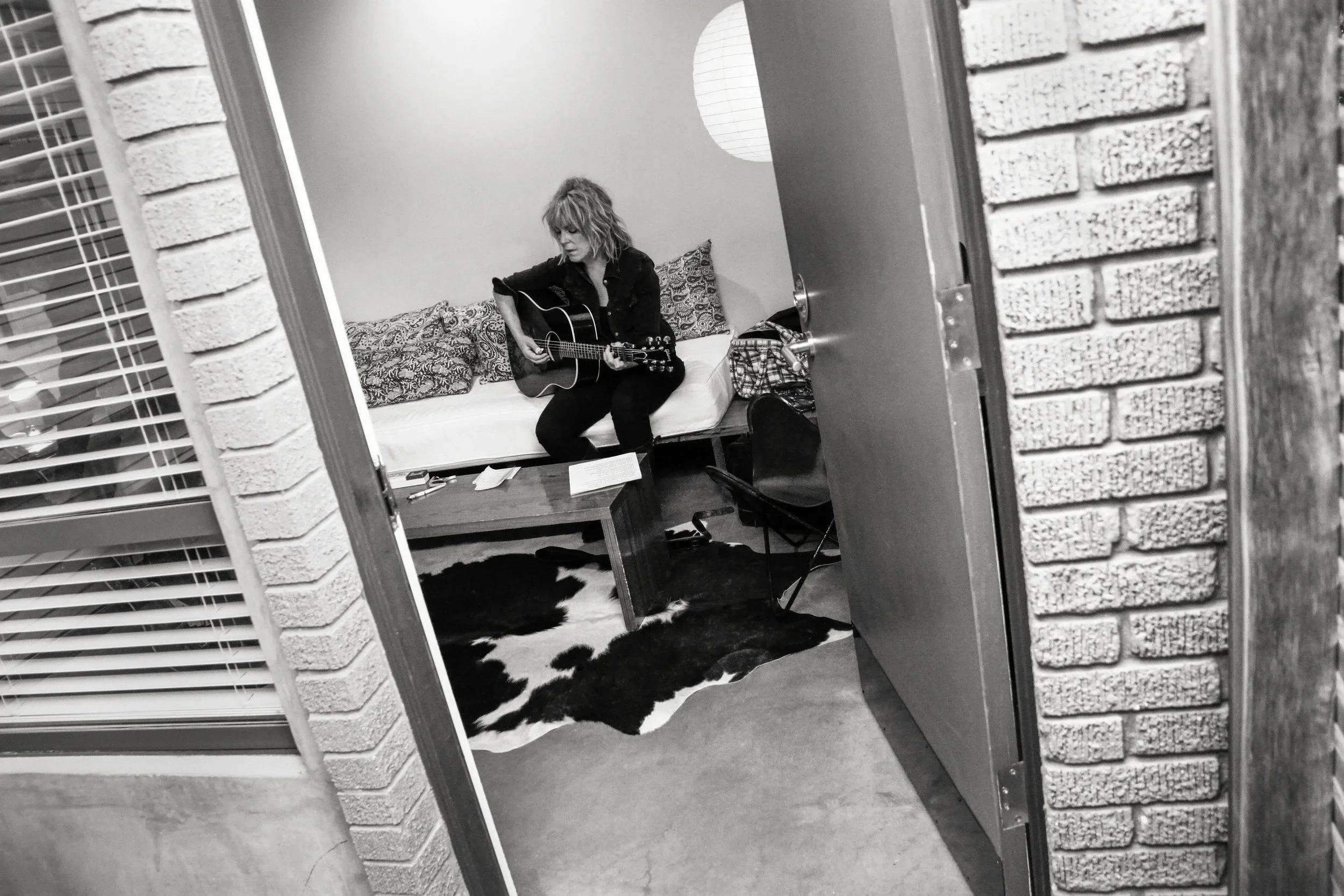 Lucinda Williams playing an acoustic guitar in a room at the Hotel San Jose, sitting on a bed, seen through an open door framed by brick walls.