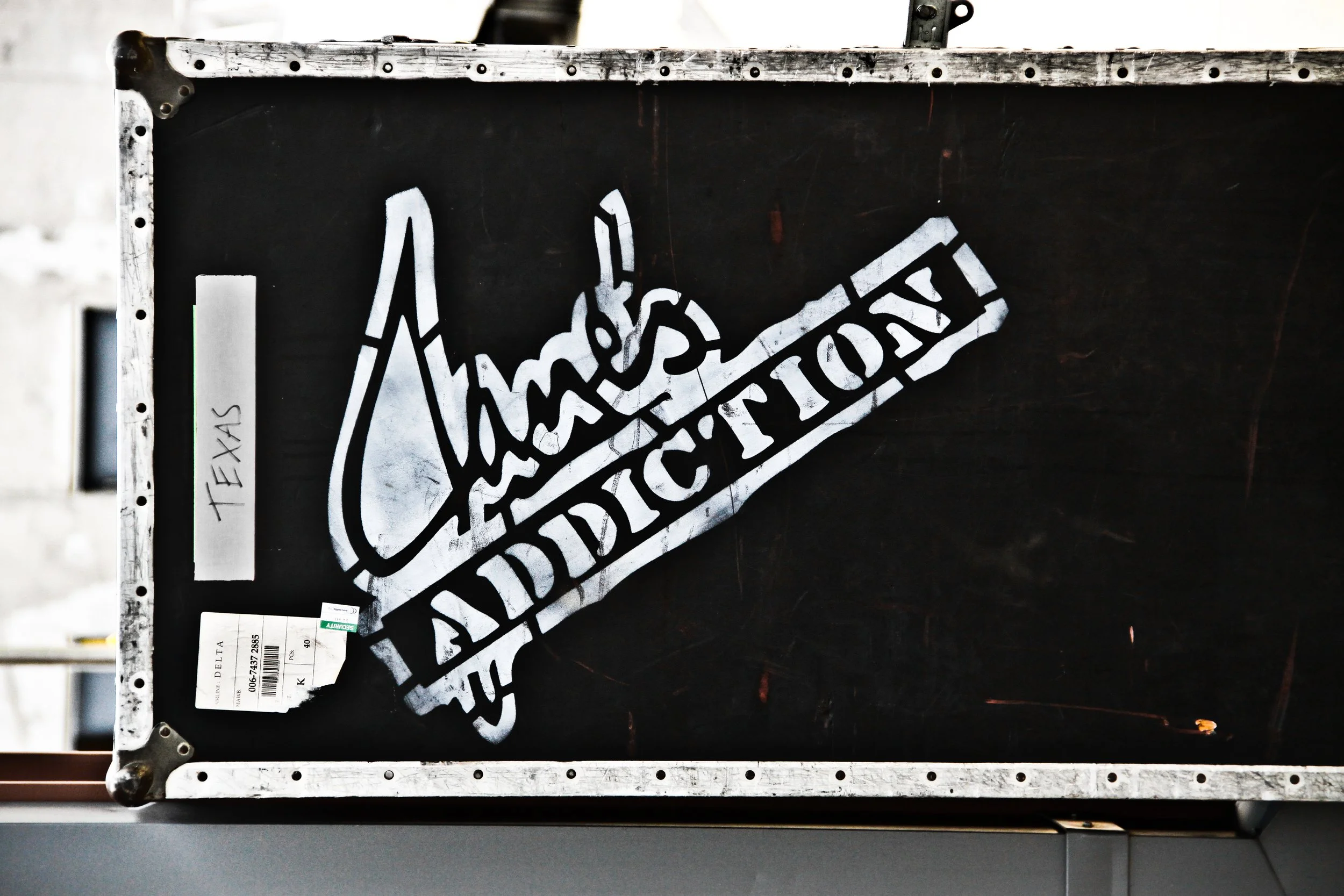 Close-up of a Jane's Addiction black crate with a white stencil design and labels, including a handwritten note that says 'TEXAS' and a partially visible green label.