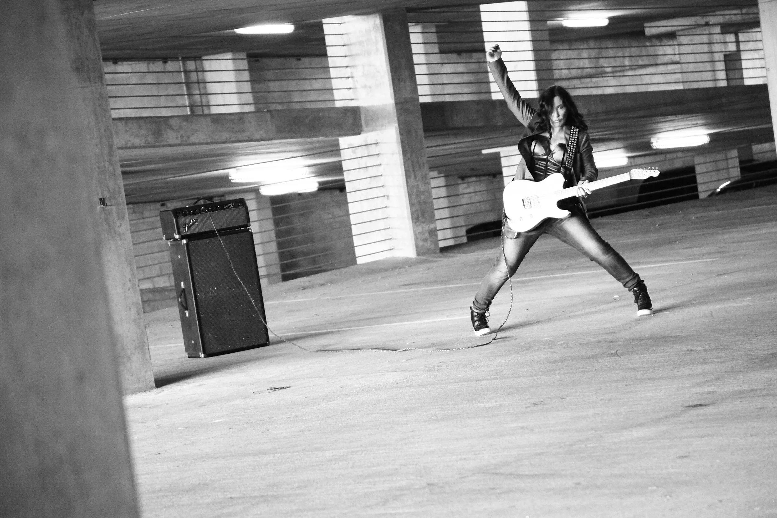 Kathy Valentine of The Go-Go's posing with an electric guitar in an urban parking garage. Black-and-white music portrait photography capturing the energy and attitude of rock and roll culture.