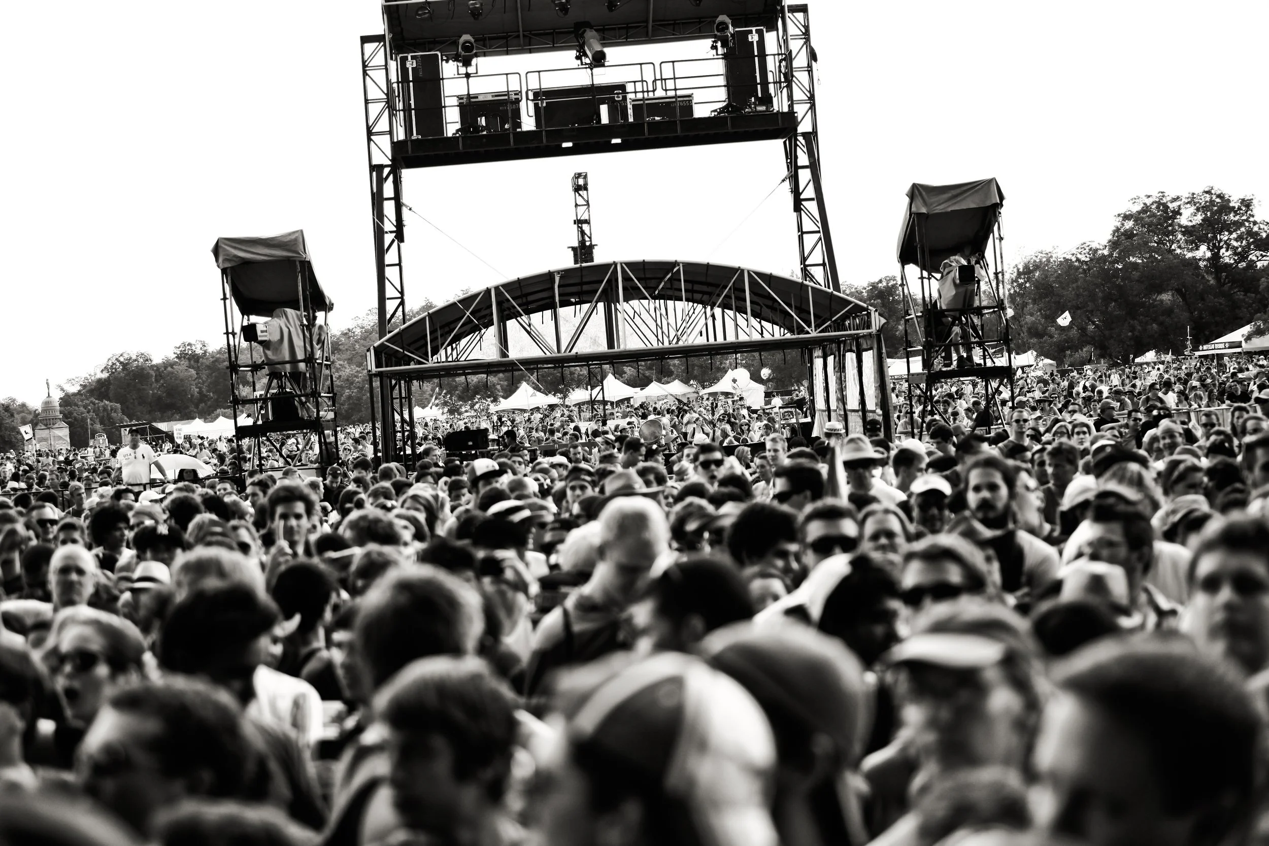 A large crowd of people attending an outdoor event or concert with a stage and equipment in the background.