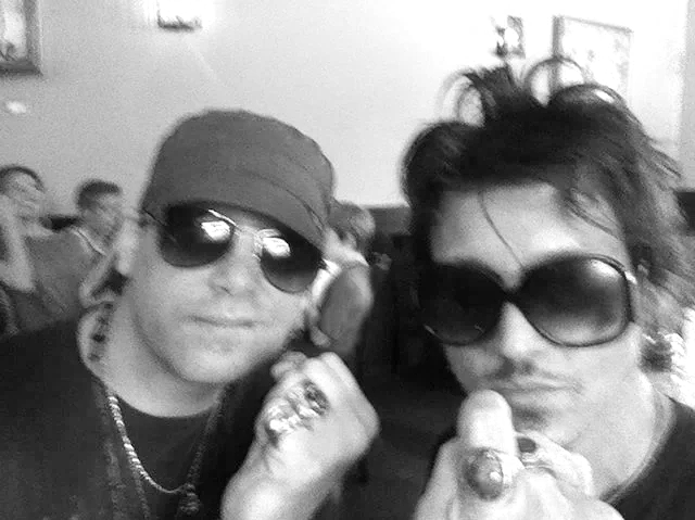 Christopher Durst and Cristo Bael wearing sunglasses, one with a green cadet hat and the other with spiky hair, posing with a mirror and a joint in a crowded cafe.