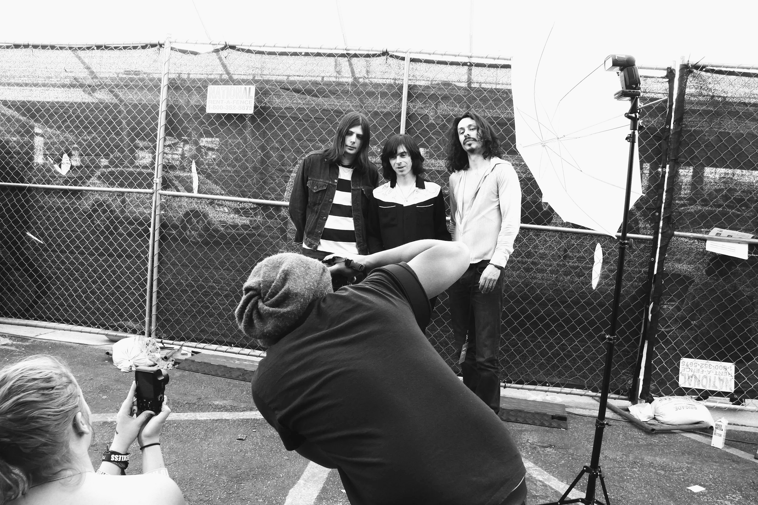 Black and white photo of Little Barrie standing behind a chain-link fence, with Christopher Durst taking a picture of them. One person is sitting on the ground, and several people in the foreground are taking photos of the scene.