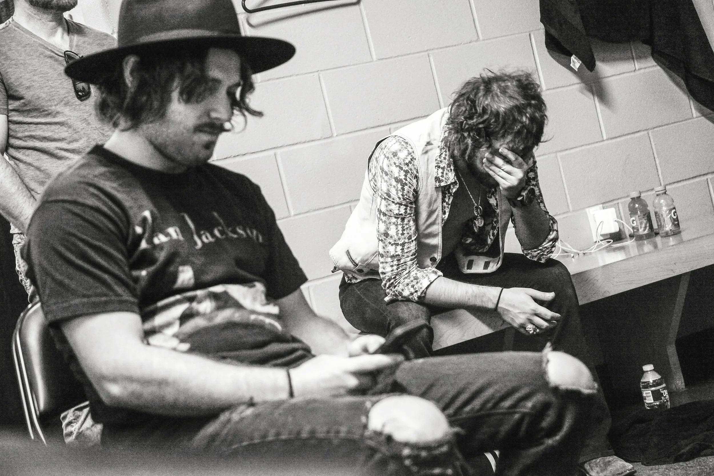 Three people sitting against a wall in a room. One man has a hat and glasses, another person, possibly a woman, with curly hair covering their face, appears distressed. The third person is partially visible, sitting next to the distressed individual. Bottles of water and bottles of Gatorade are on a table nearby.