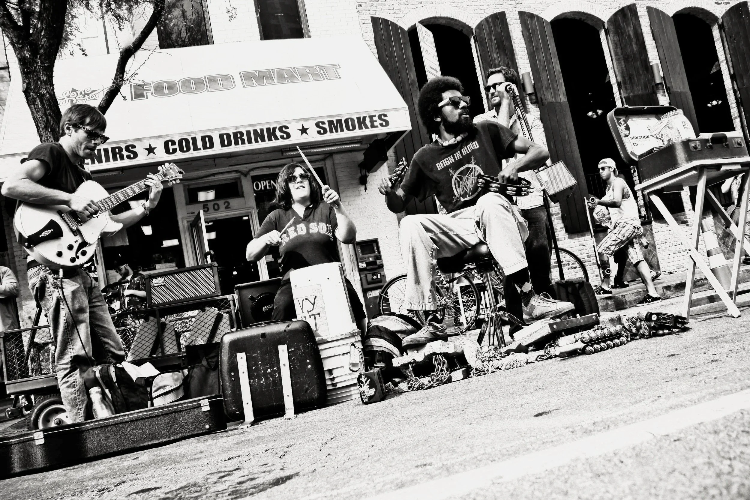 Street music band performing outdoors in front of a building with a food mart sign, surrounded by various objects and a few pedestrians.
