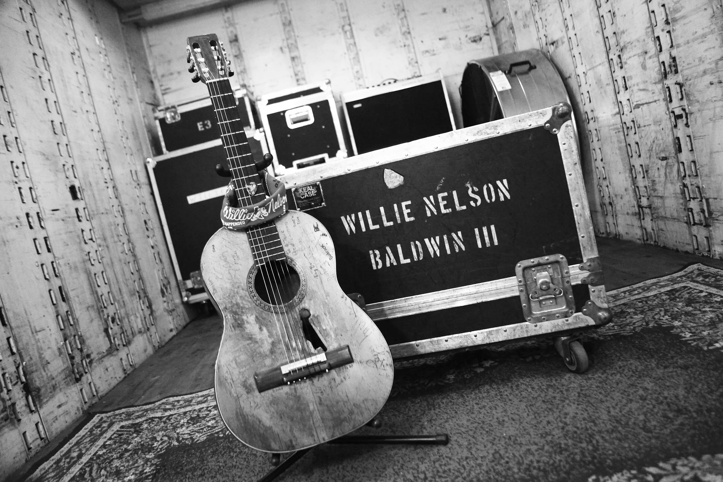 A black and white photograph of a wooden floor and wall, with a vintage style guitar leaning against a large utility case labeled "Willie Nelson Baldwin, TX," surrounded by other cases, a rug, and a snare drum.