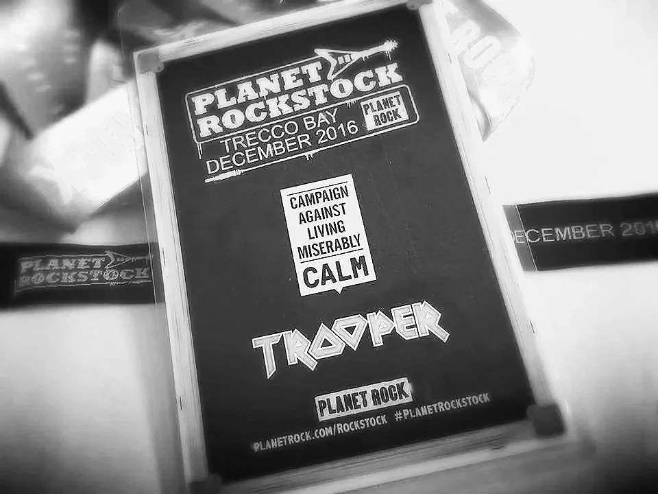 Black and white photo of a card or brochure with text about the band Planet Rockstock at Trecco Bay, December 2016, promoting a campaign against living miserably with a message to stay calm, featuring the word "Trapper" and the Planet Rock website address.
