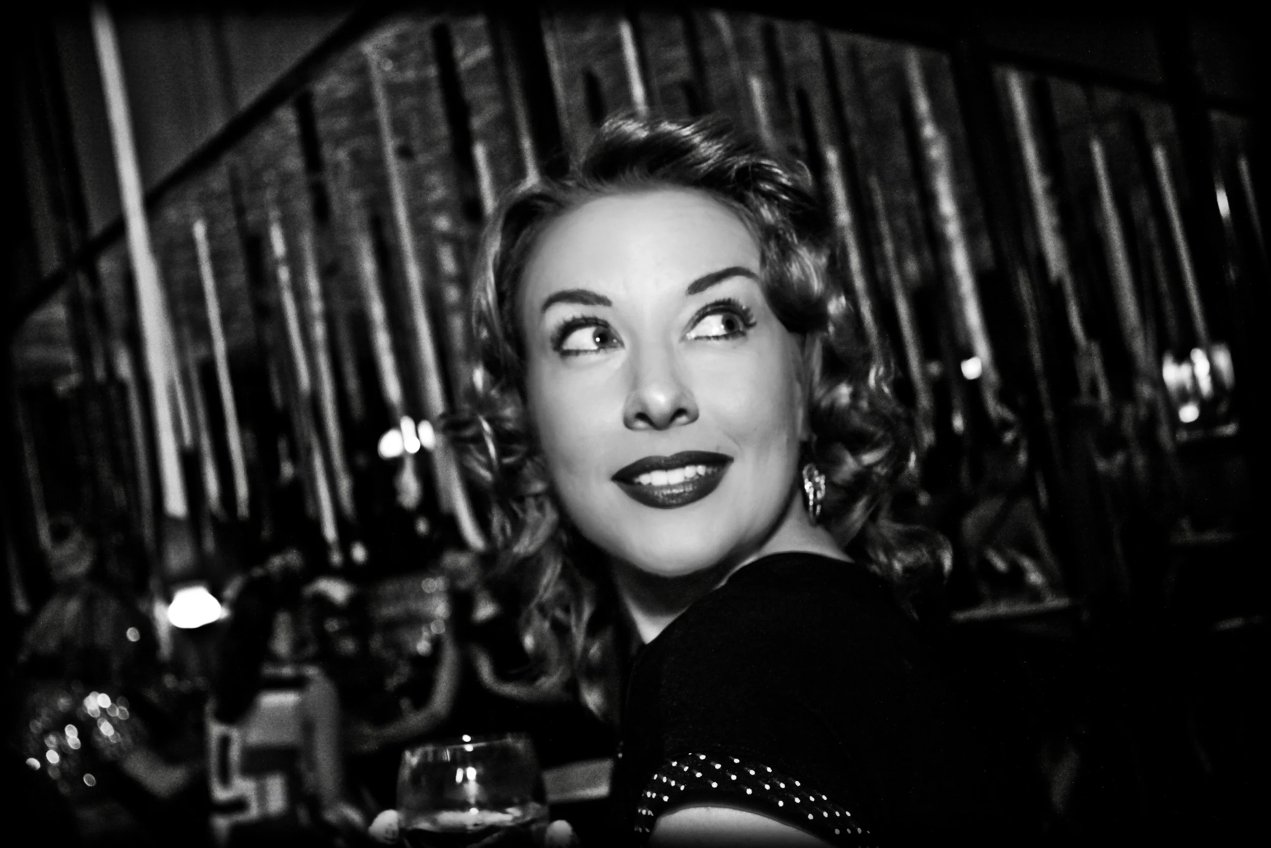 A woman with curly hair and vintage style makeup, smiling and looking to the side, standing in front of a background with vertical bars.