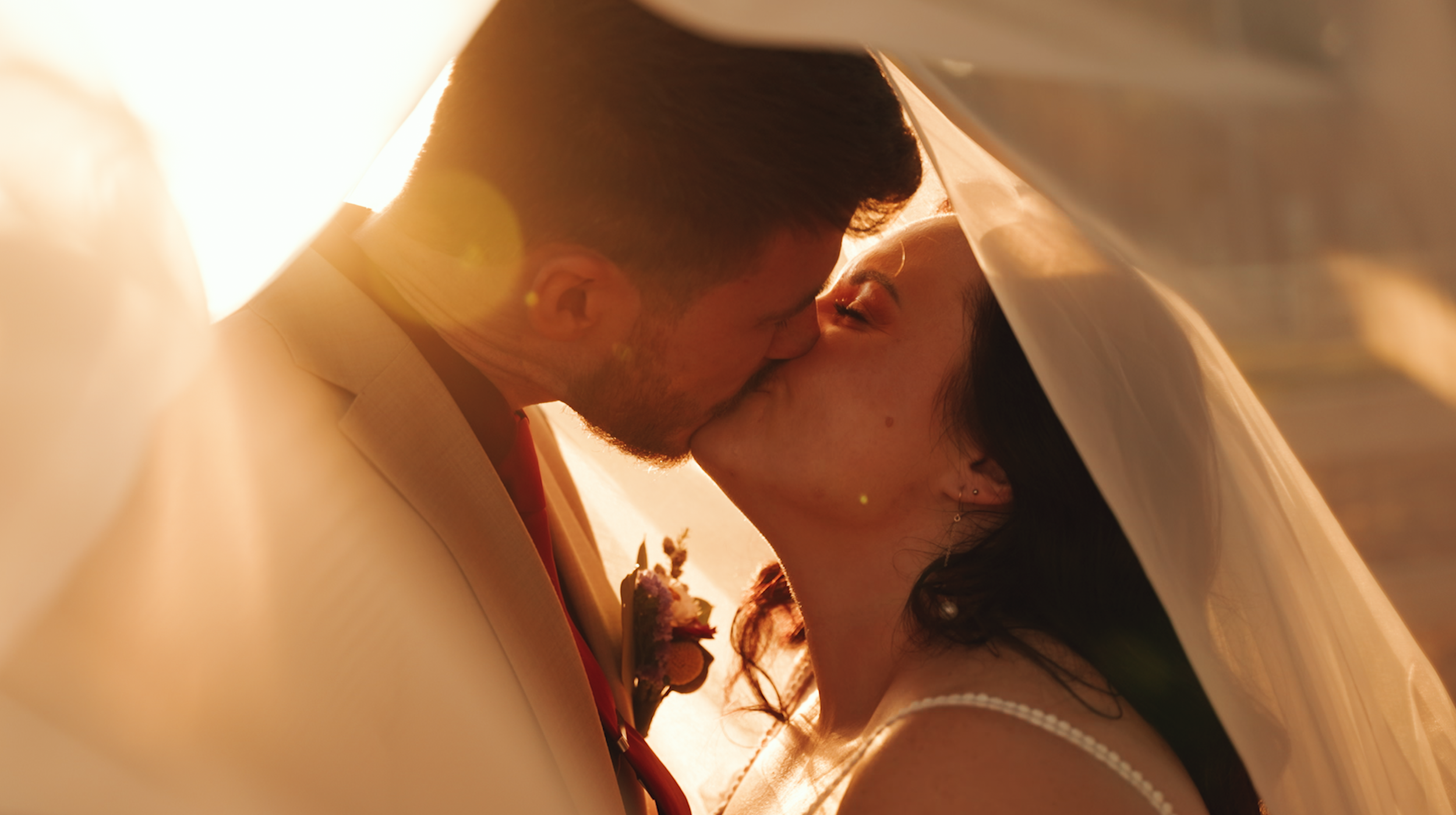 A couple sharing a kiss under a wedding veil during sunset.