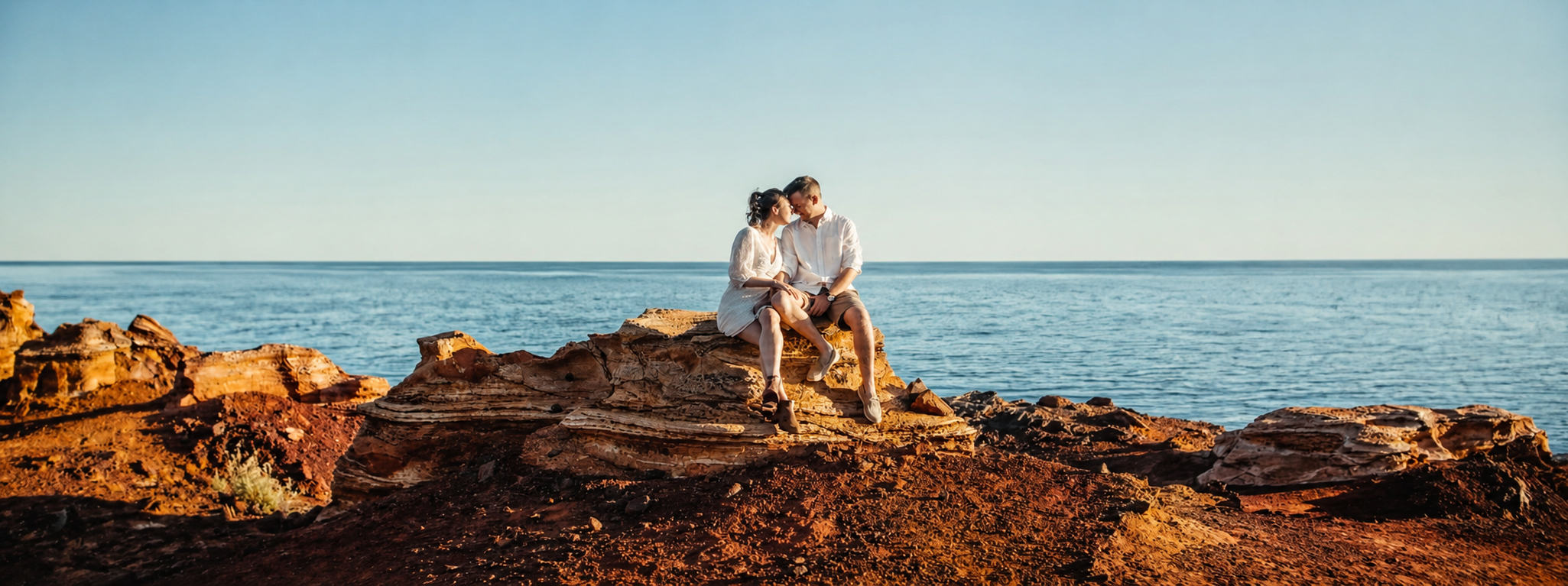 A couple sitting on a large rock formation by the ocean, close to each other, with the sea and clear sky in the background.