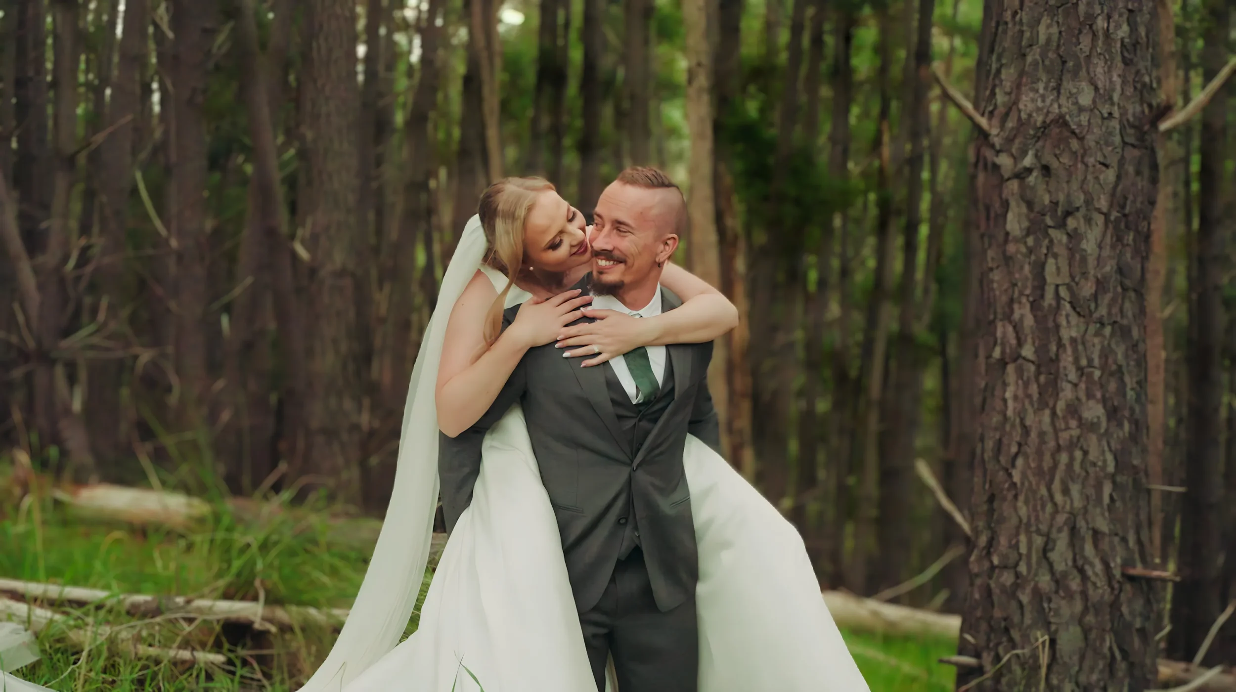 A bride and groom in wedding attire sharing a joyful moment in a forest, with the bride hugging the groom from behind, trees and greenery in the background.