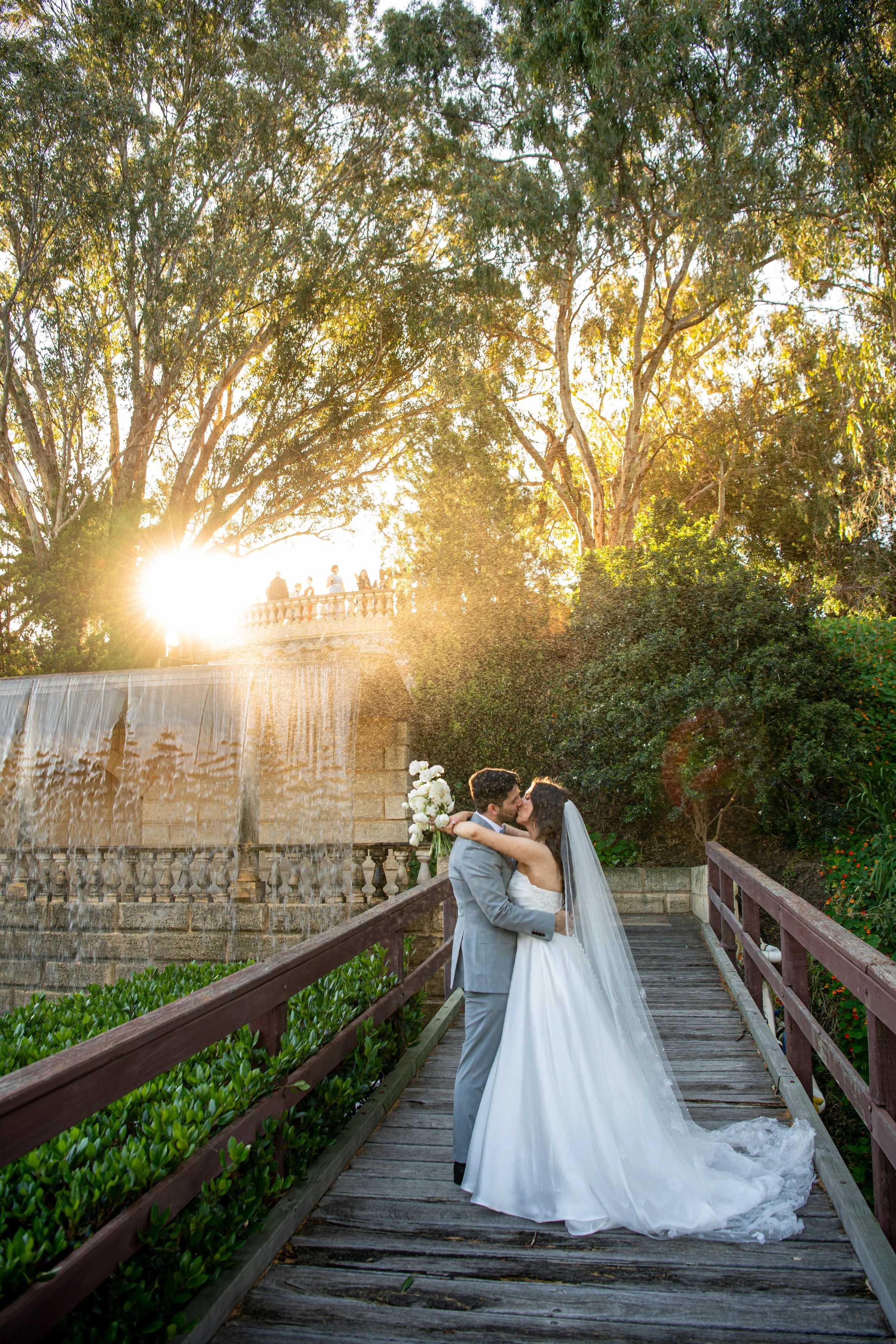 A bride and groom kissing on a wooden bridge surrounded by lush greenery and trees, with the sun setting in the background and a fountain nearby.