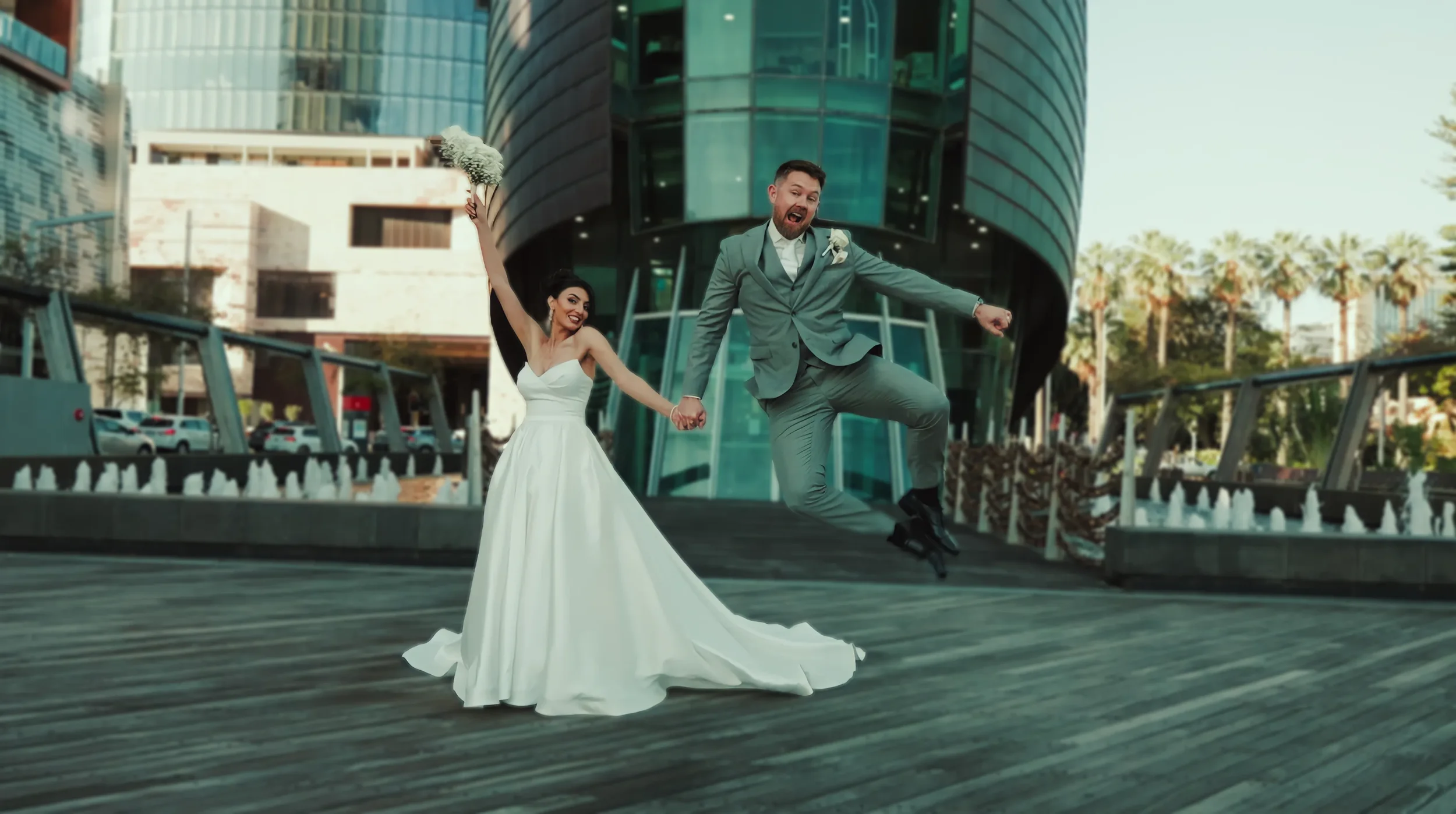 A bride and groom celebrating outside a modern glass building, holding hands, with the groom jumping in the air wearing a gray suit and the bride smiling in a white wedding dress, holding a bouquet of flowers.