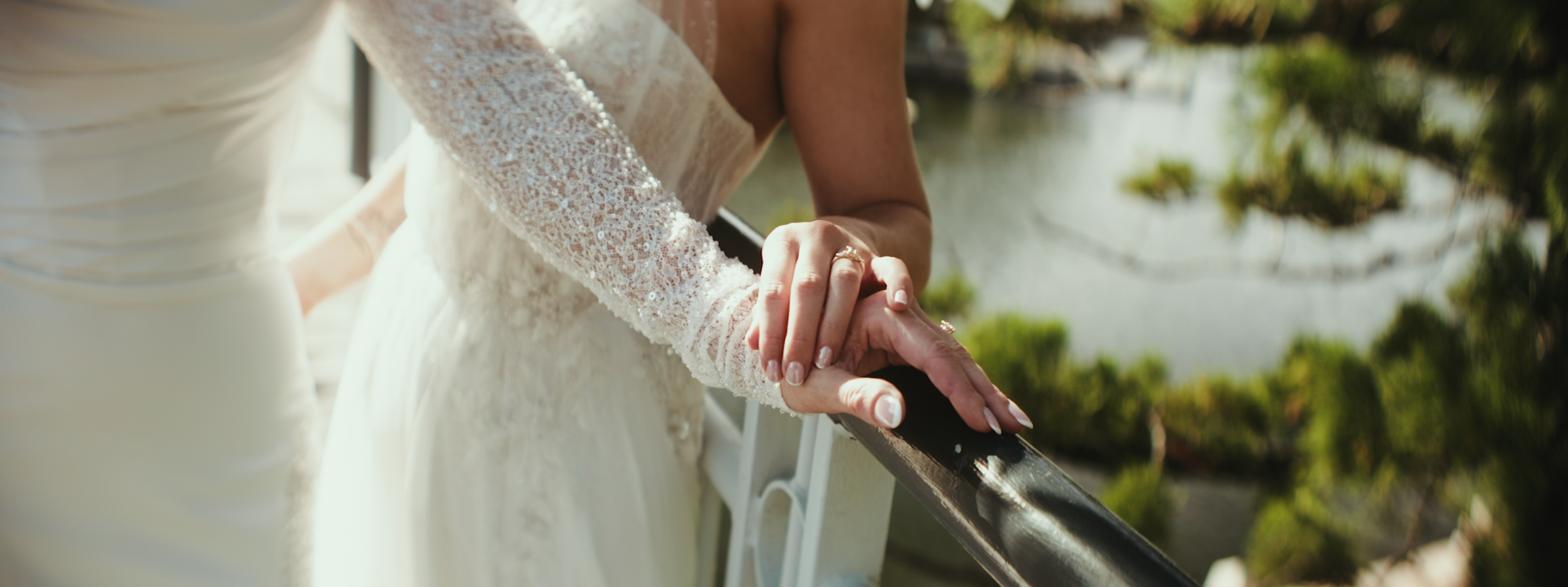 A bride in a lace wedding dress holding hands with her partner during a wedding ceremony outdoors, with water and green trees in the background.