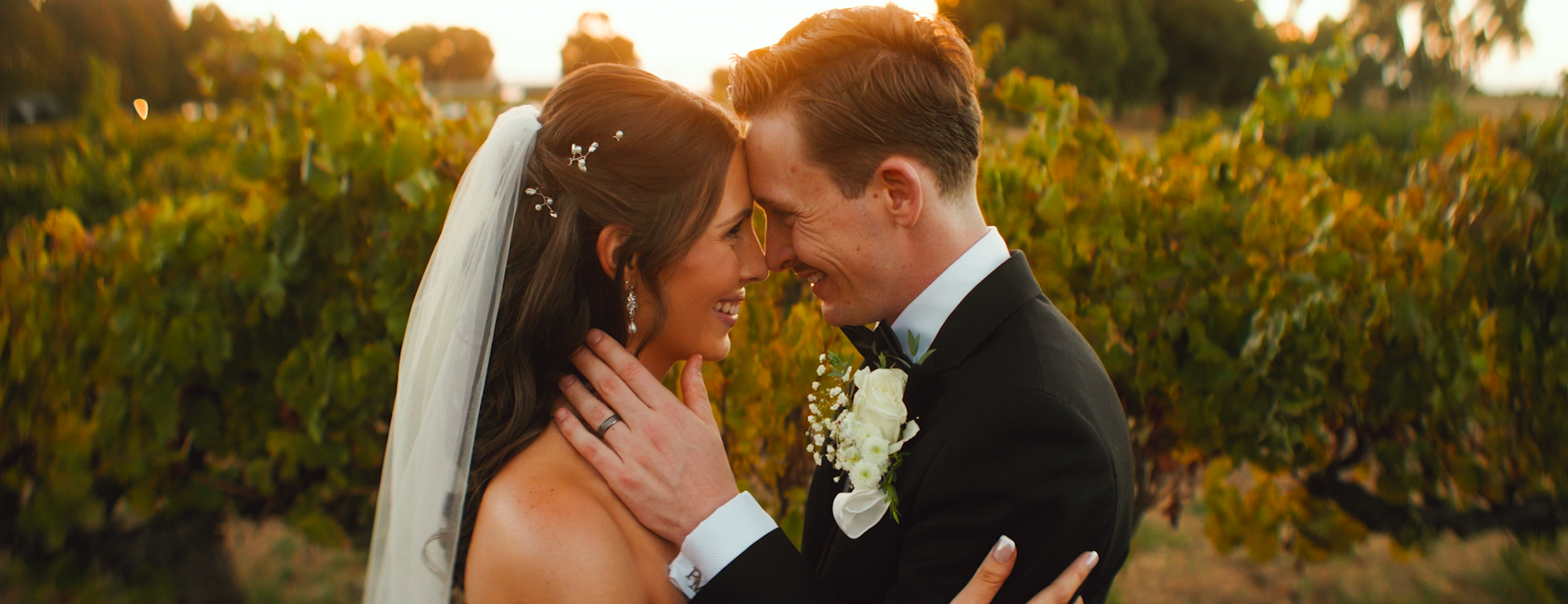 A bride and groom sharing a close, joyful moment outdoors during sunset, touching foreheads with eyes closed, surrounded by green and yellow foliage.