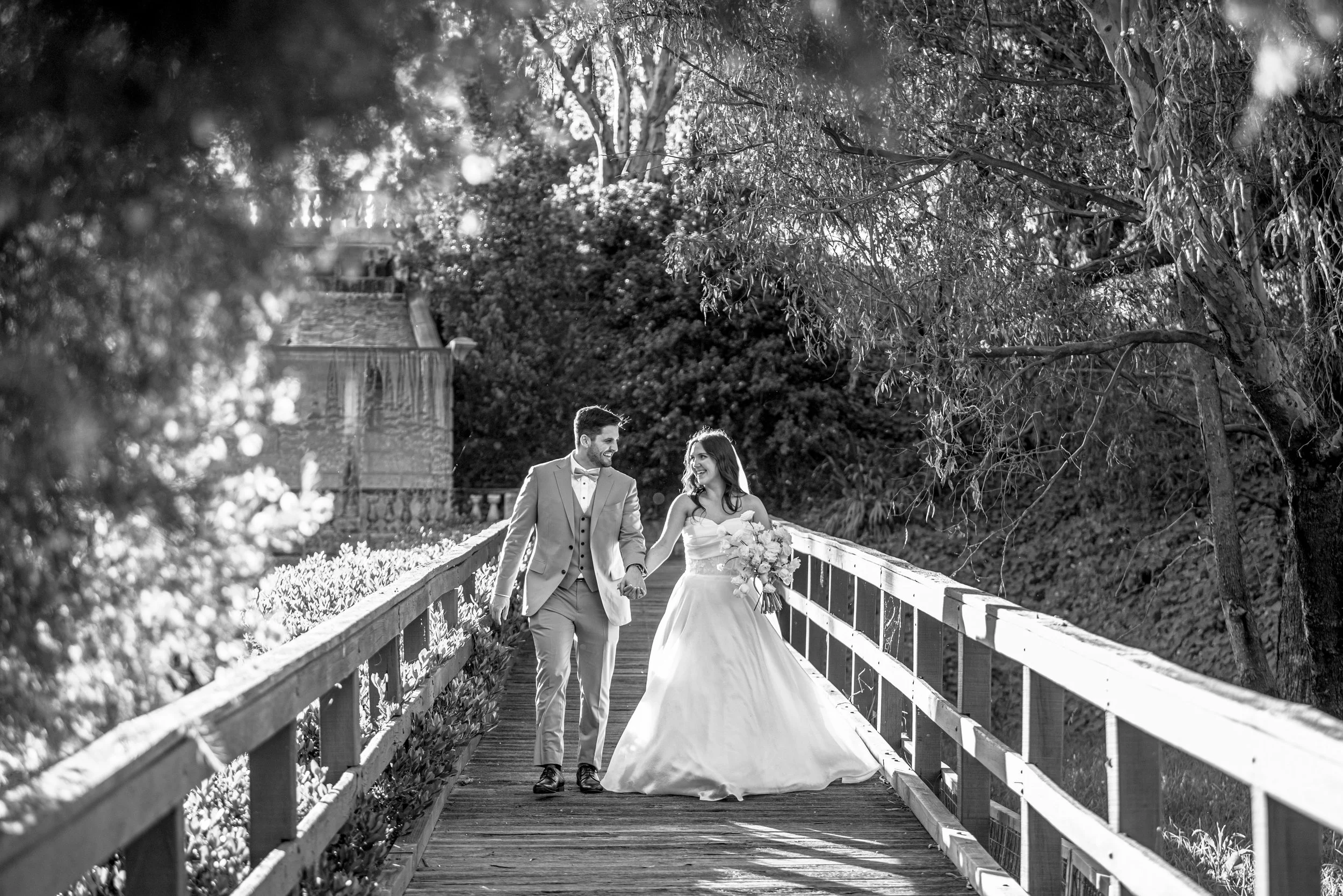 A happy newlywed couple holding hands and walking on a footbridge in a park with trees, during daytime, black and white photo.