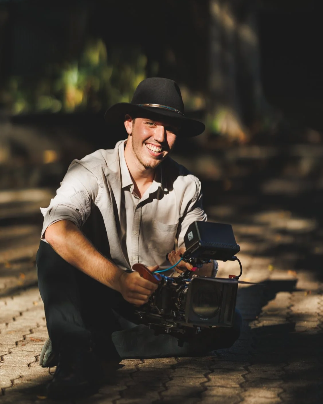 A man wearing a black wide-brimmed hat and light-colored shirt, smiling and kneeling on a cobblestone path while holding a professional video camera.