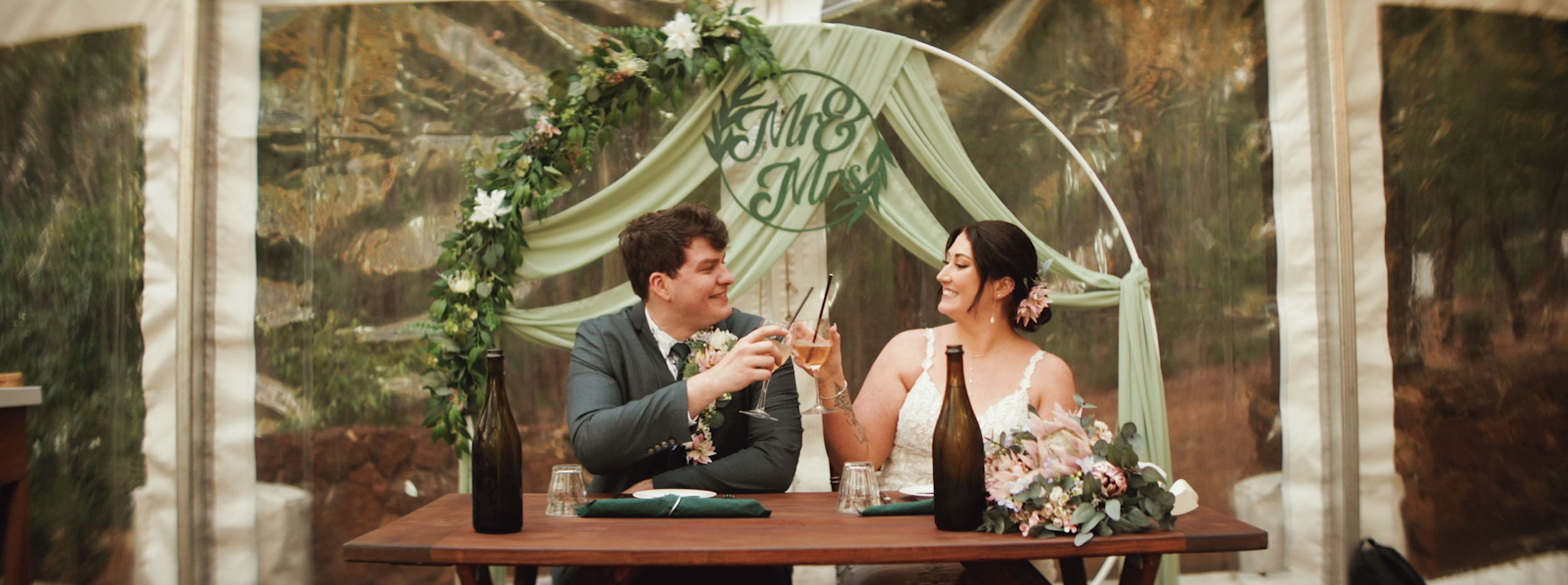 A newlywed couple celebrating at their wedding reception, sitting at a table with drinks, surrounded by wedding decor and greenery, smiling at each other.