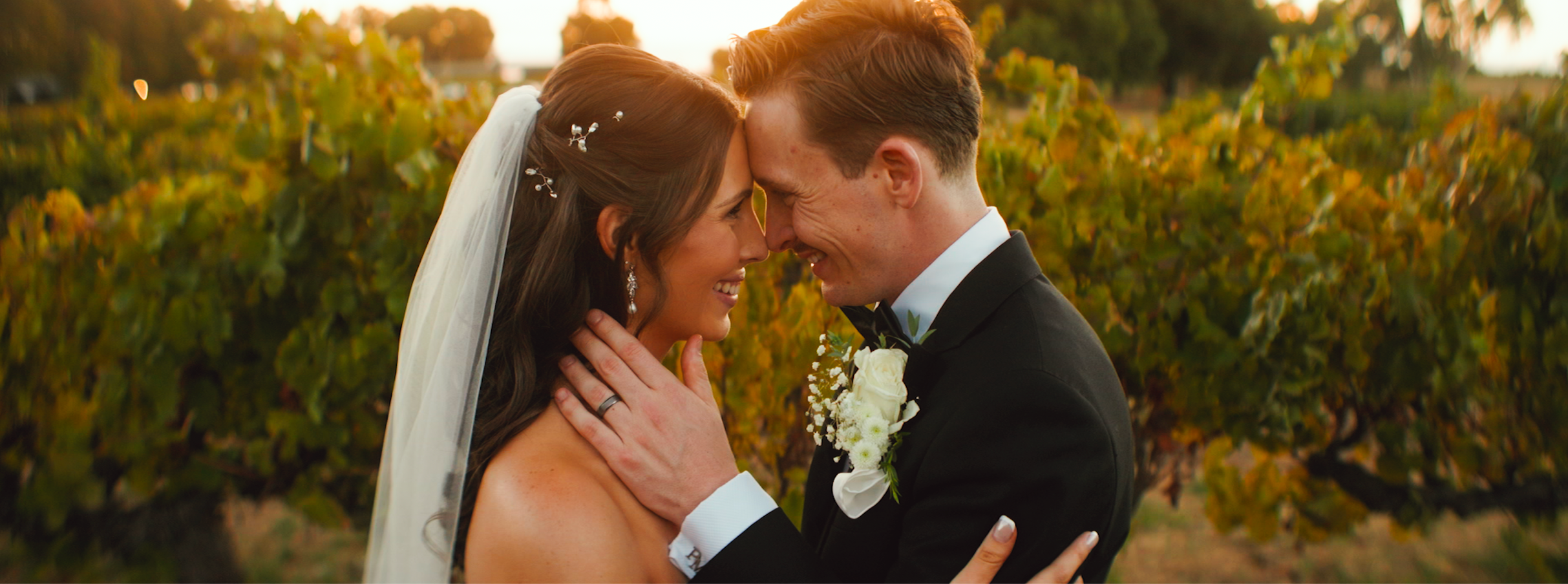 A bride and groom sharing a close, joyful moment outdoors during sunset, touching foreheads with eyes closed, surrounded by green and yellow foliage.