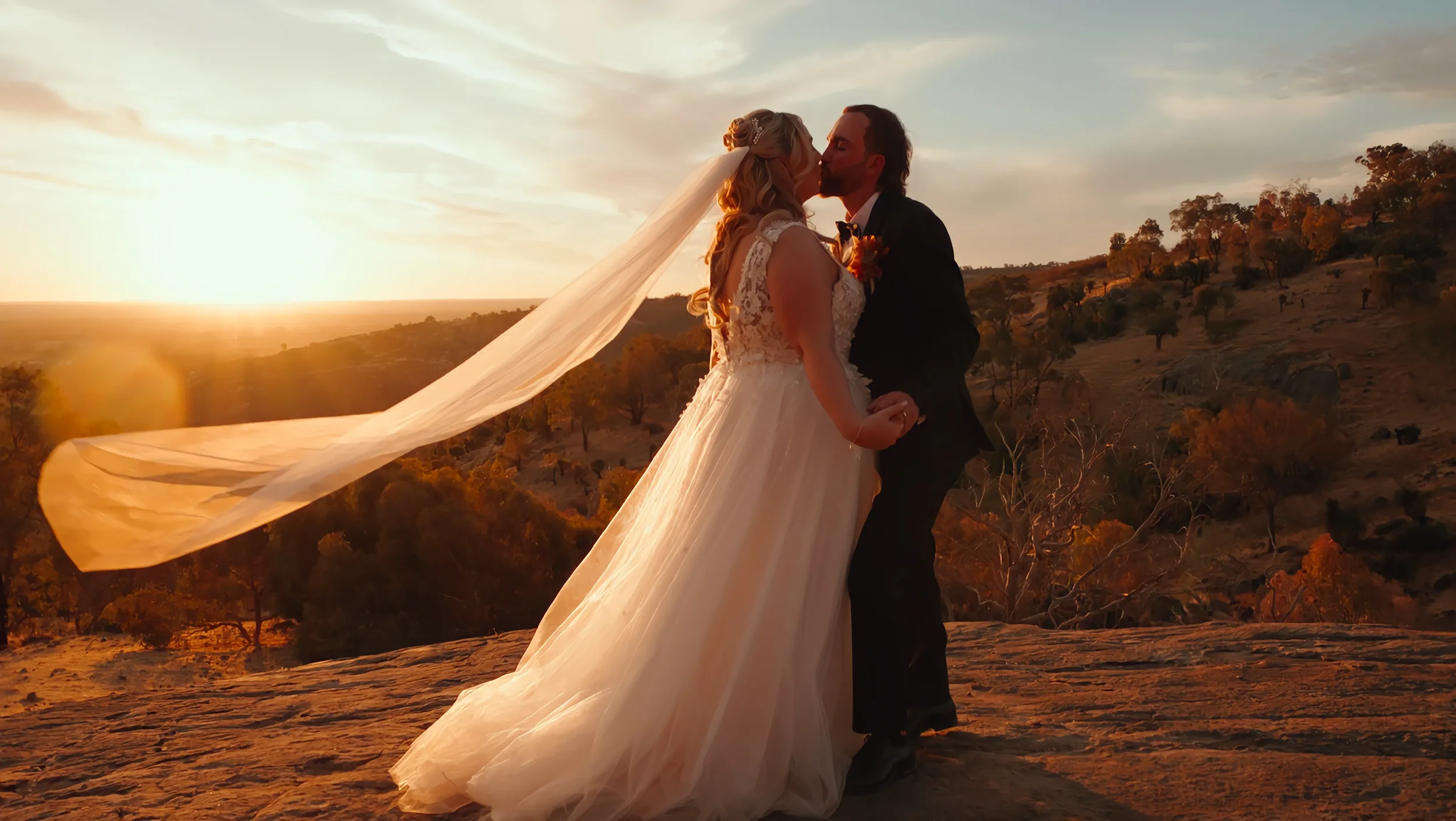 A bride and groom sharing a kiss during sunset outdoors, with trees and hills in the background.