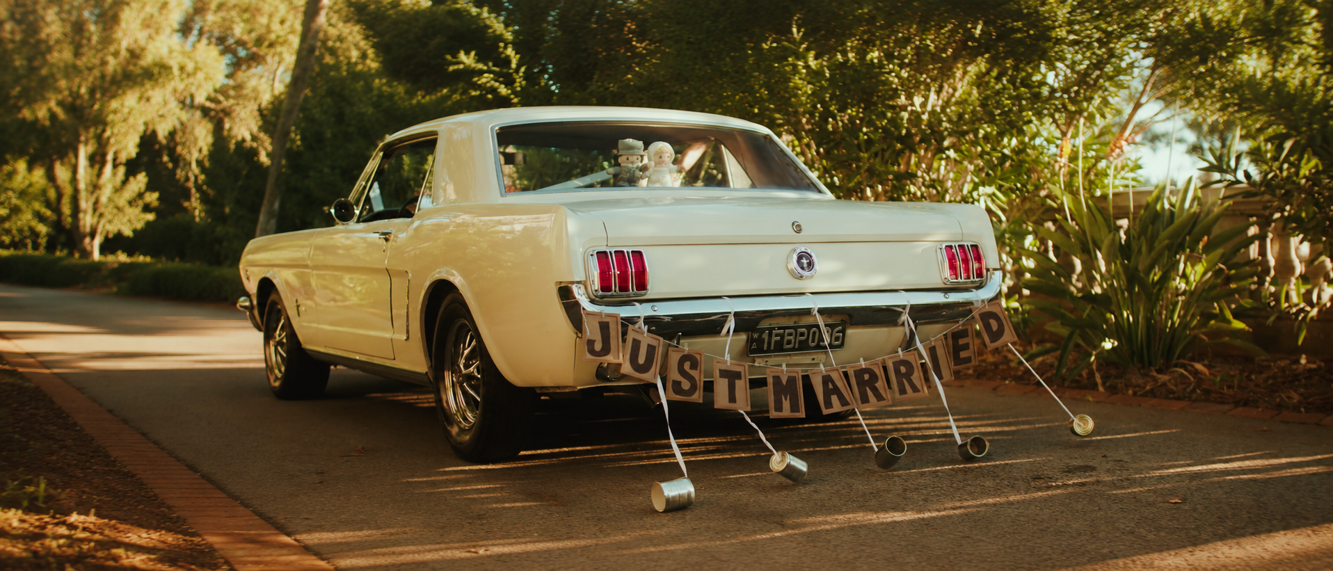 A vintage white car decorated with a 'Just Married' sign on the back, parked on a paved road surrounded by trees and greenery during sunset.