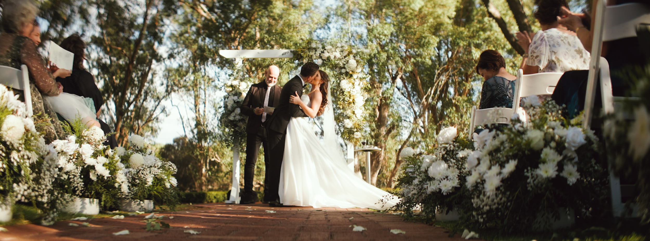 A couple exchanging vows and kissing at an outdoor wedding ceremony surrounded by floral decorations and guests seated on white chairs.