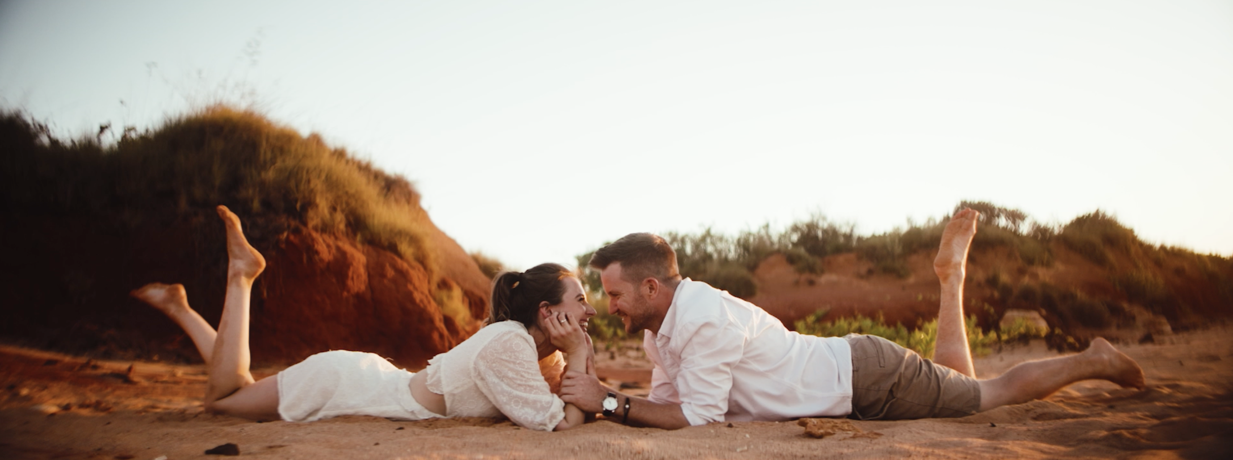 A couple lying on the sandy beach, facing each other, smiling and laughing during sunset with dunes and bushes in the background.