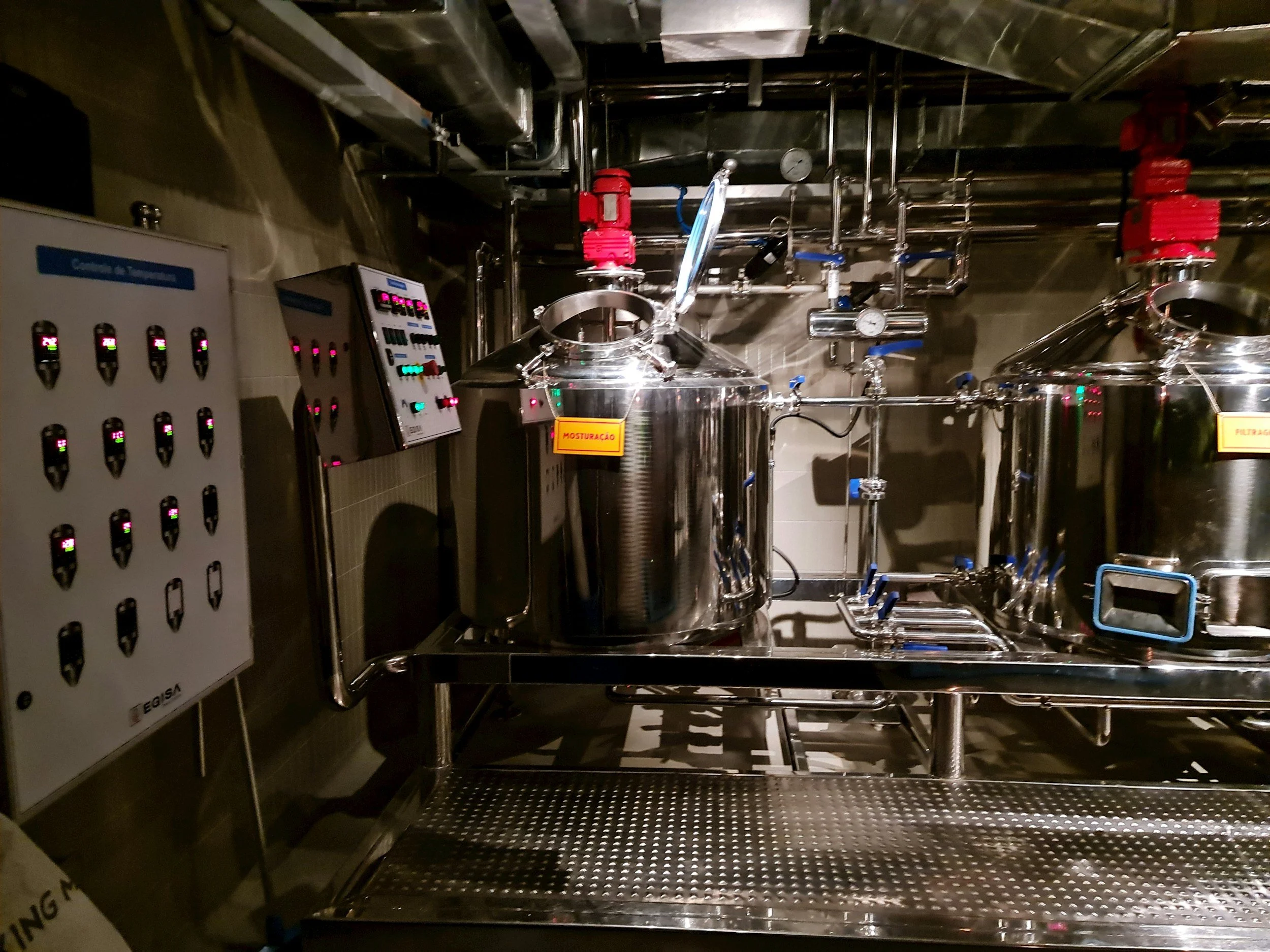Industrial equipment in a dimly lit room, including stainless steel tanks, pipes, gauges, and control panels, used for manufacturing or processing.